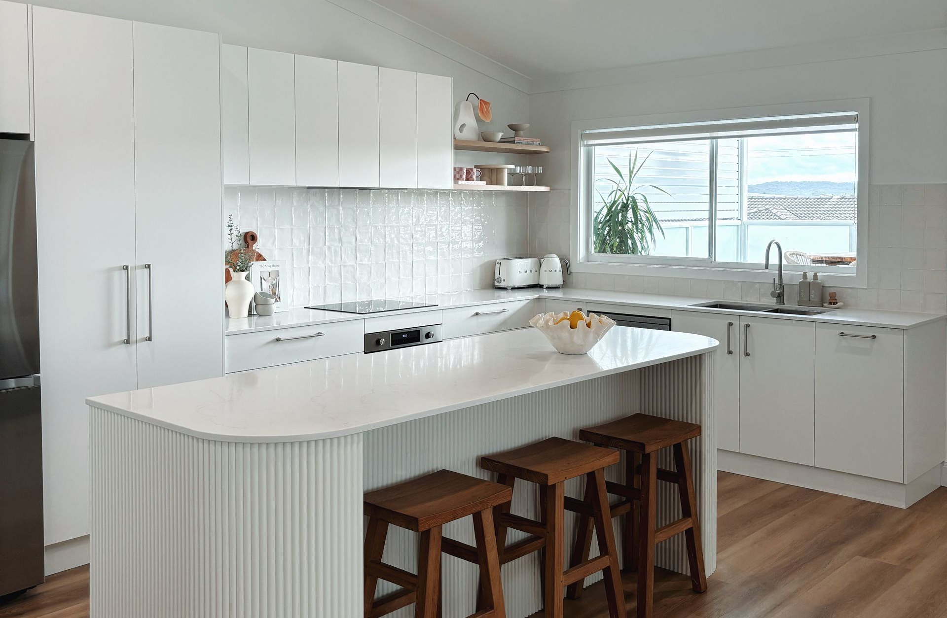 White kitchen with a curved island, wooden stools, and a window overlooking a view.
