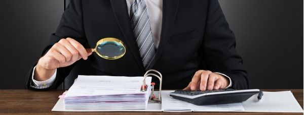 A man is sitting at a desk with a magnifying glass scrutinizing a Medicaid application.