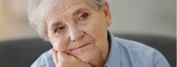 A depressed elderly woman is sitting on a couch with her hand on her chin.