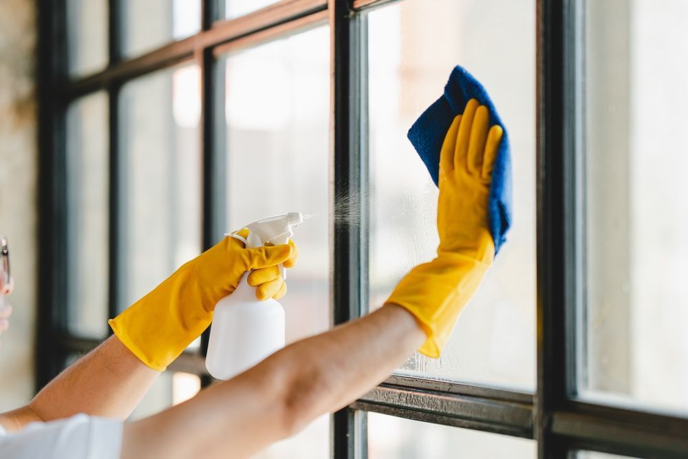 Person wearing yellow gloves sprays and wipes a window with a blue cloth.