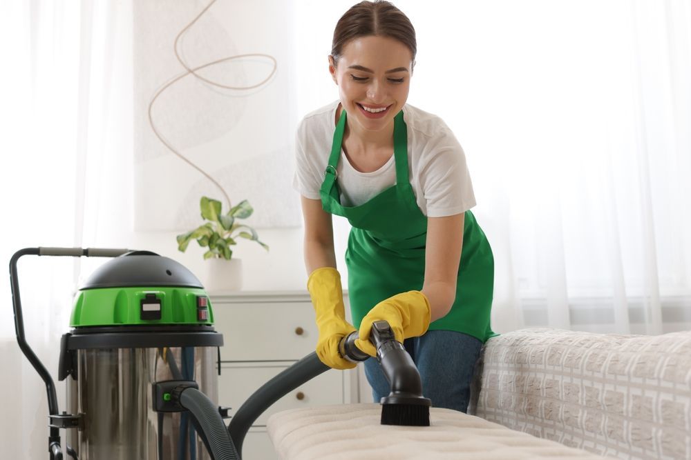 Woman vacuuming a couch with a commercial vacuum cleaner, smiling in a brightly lit room.