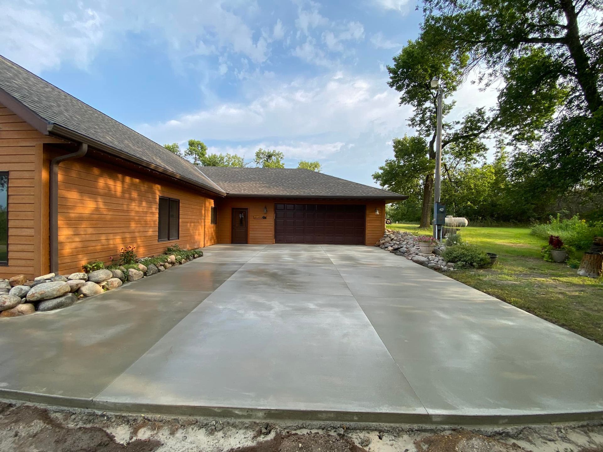 A newly poured concrete driveway leads to the garage of a brown-sided house surrounded by trees and decorative rocks.