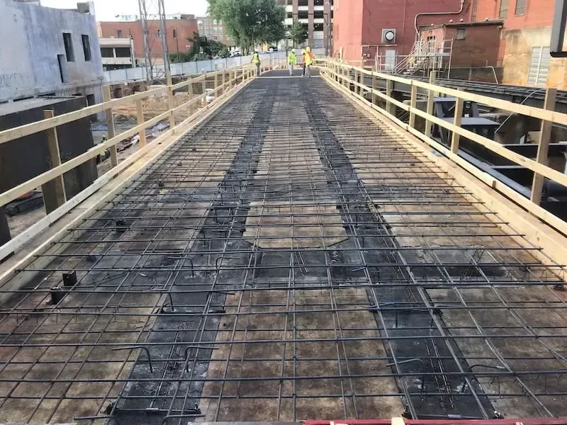 A construction site view of a bridge deck with exposed metal rebar reinforcement and temporary wooden safety railings.