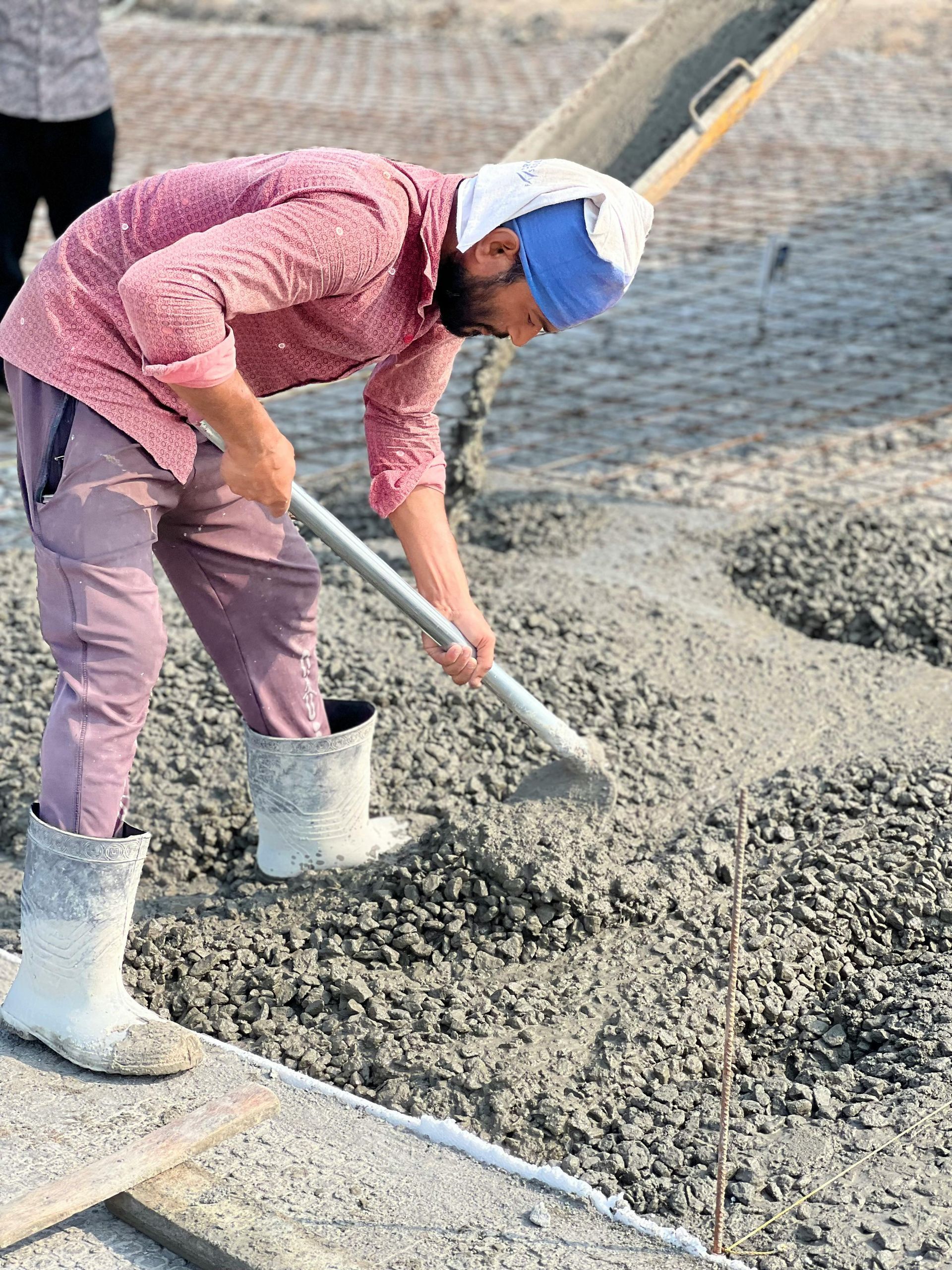 A person in a pink shirt and white rubber boots spreads wet concrete on a construction site with a metal tool.
