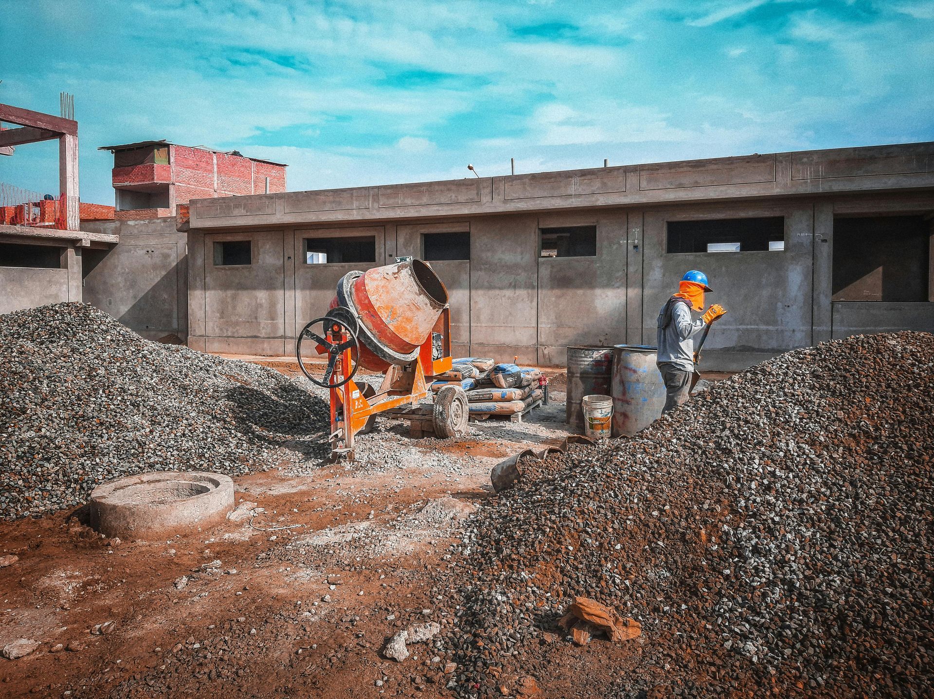 A construction worker in safety gear stands near a concrete mixer and piles of gravel at an active building site.