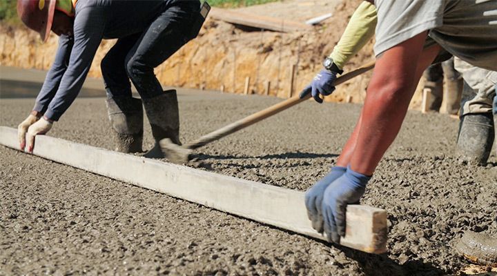 Construction workers use a long wooden board to level freshly poured concrete at a job site.