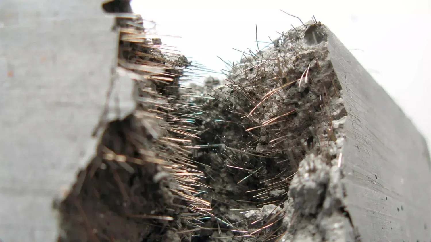 A close-up view of a fractured concrete beam revealing exposed, thin metal fibers used for reinforcement within the material.