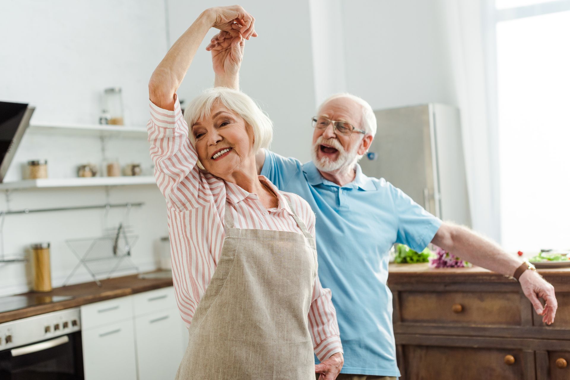 Elderly couple dances joyfully in a bright kitchen, woman in apron and man with open mouth.