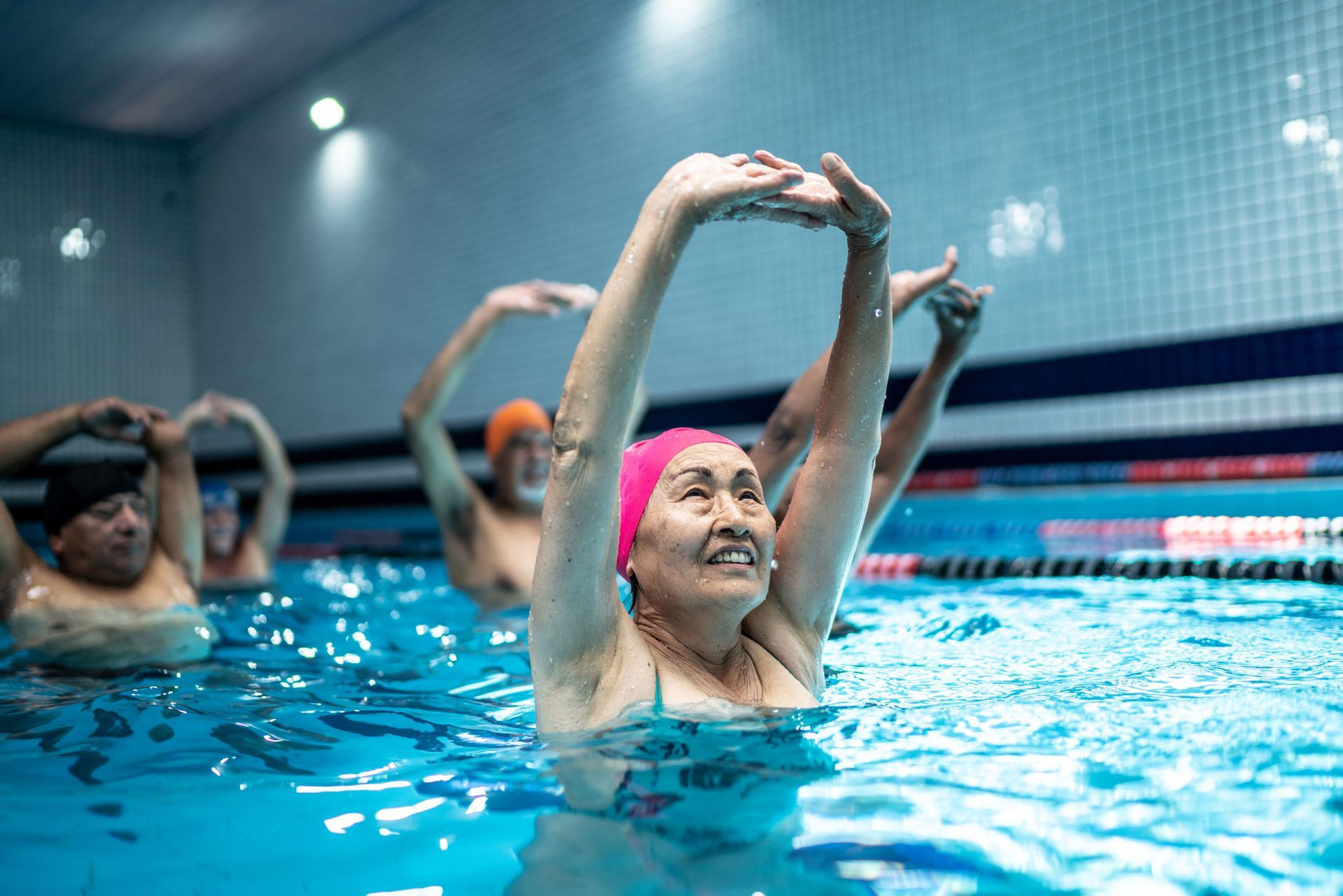 People in a pool doing water aerobics: older woman with pink cap smiles, arms raised.