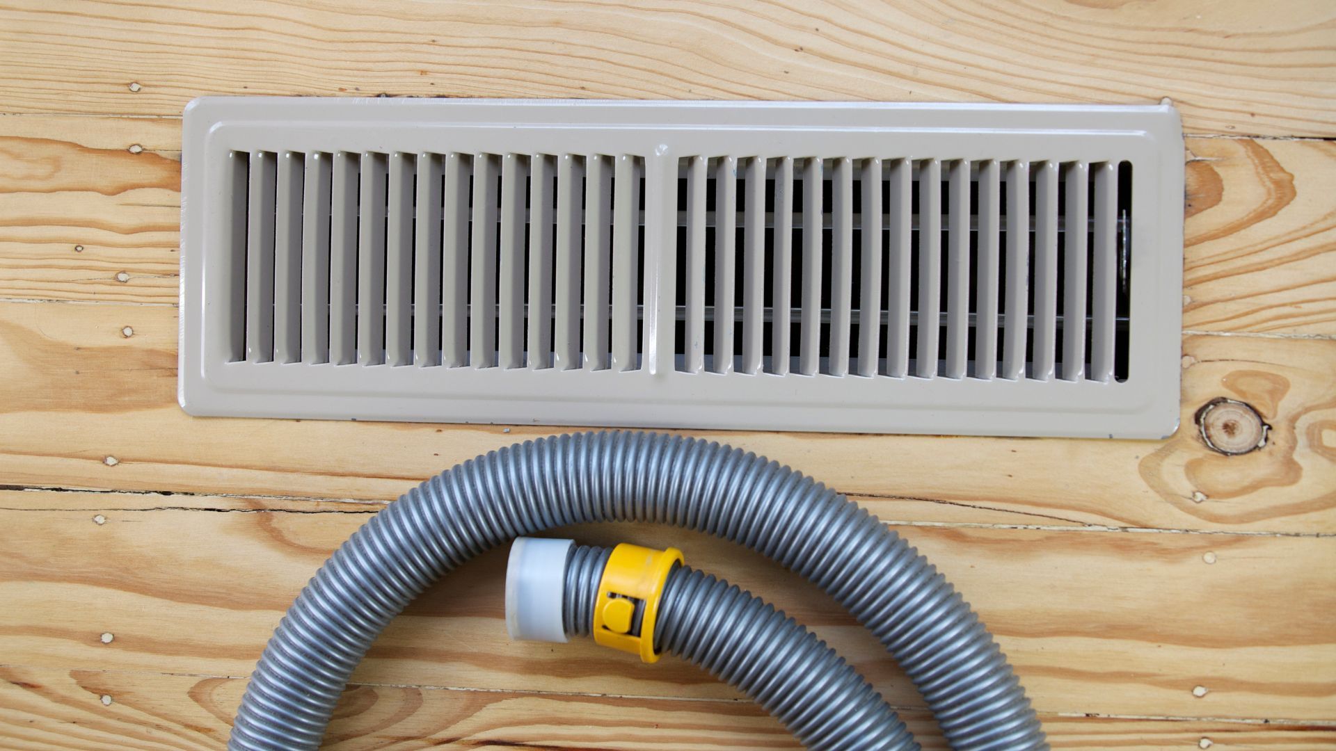 A vacuum hose is attached to a vent on a wooden floor.