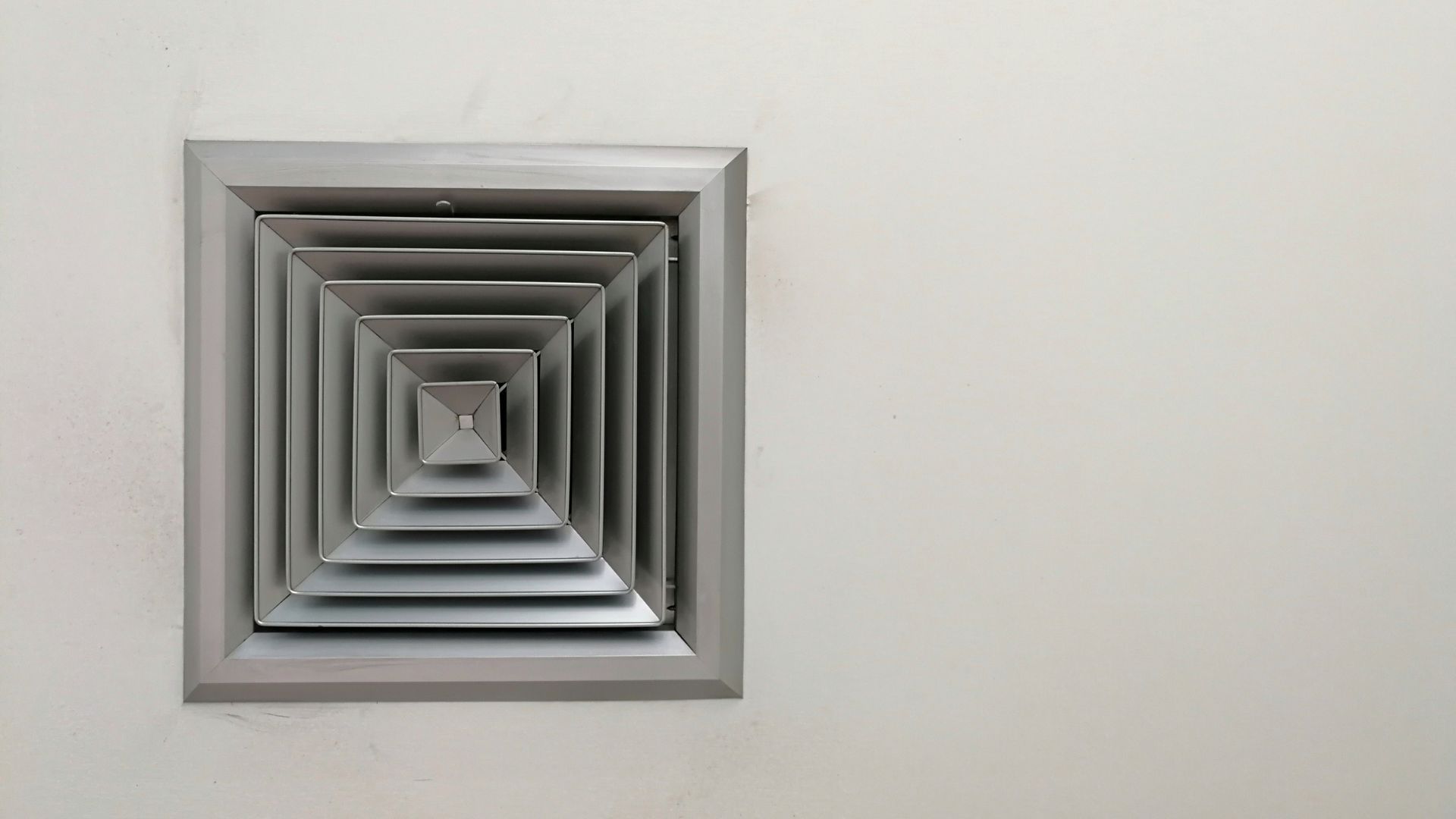 A square air vent is hanging from the ceiling on a white wall.