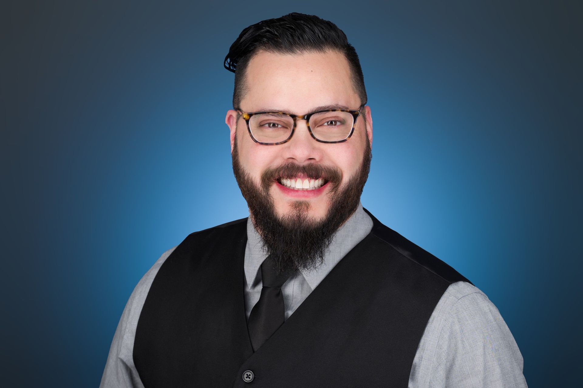 A person with a beard and glasses, wearing a light gray shirt and dark vest, smiling against a blue background.