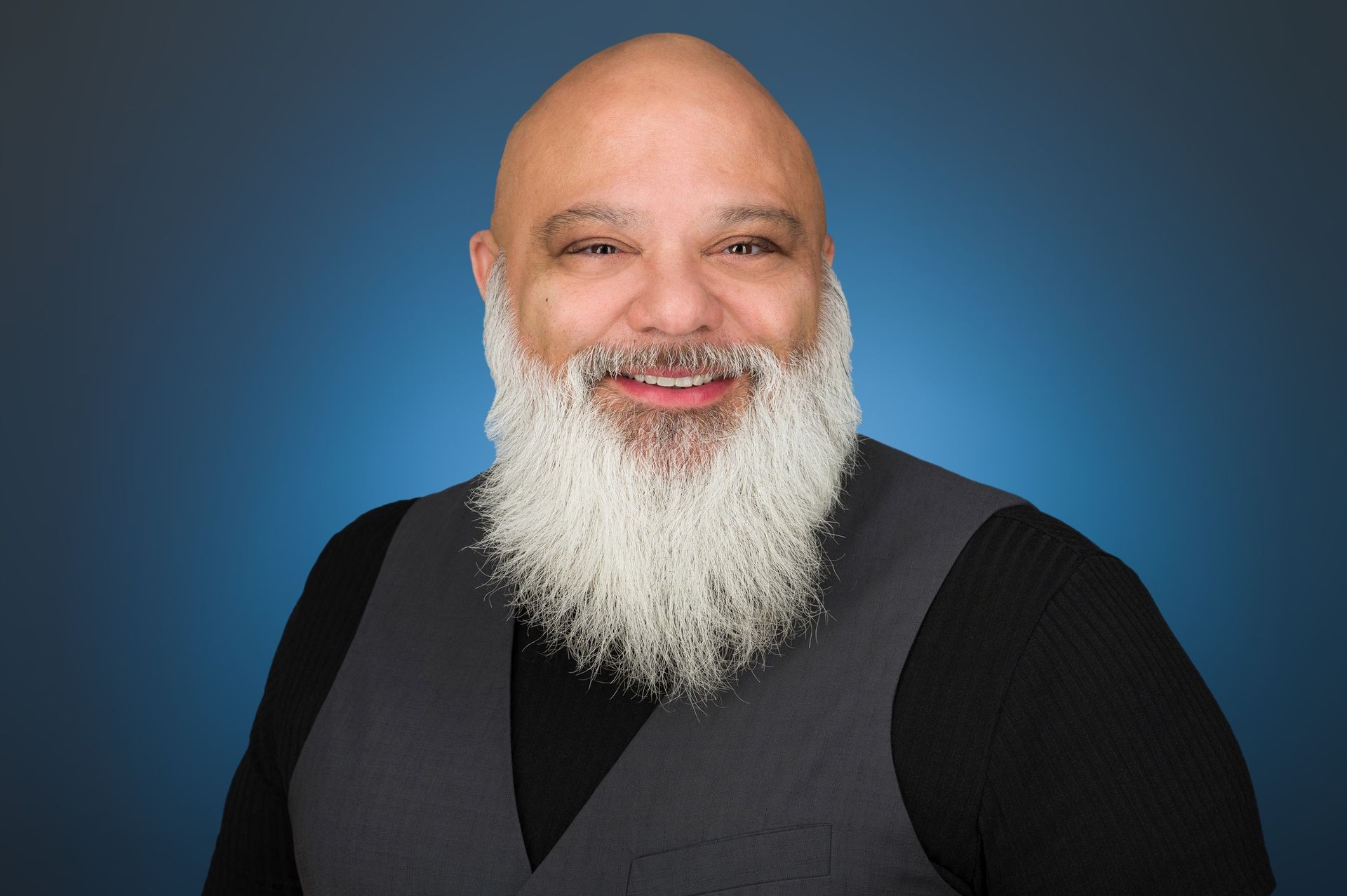 A smiling person with a long white beard, wearing a black shirt and grey vest against a blue background.