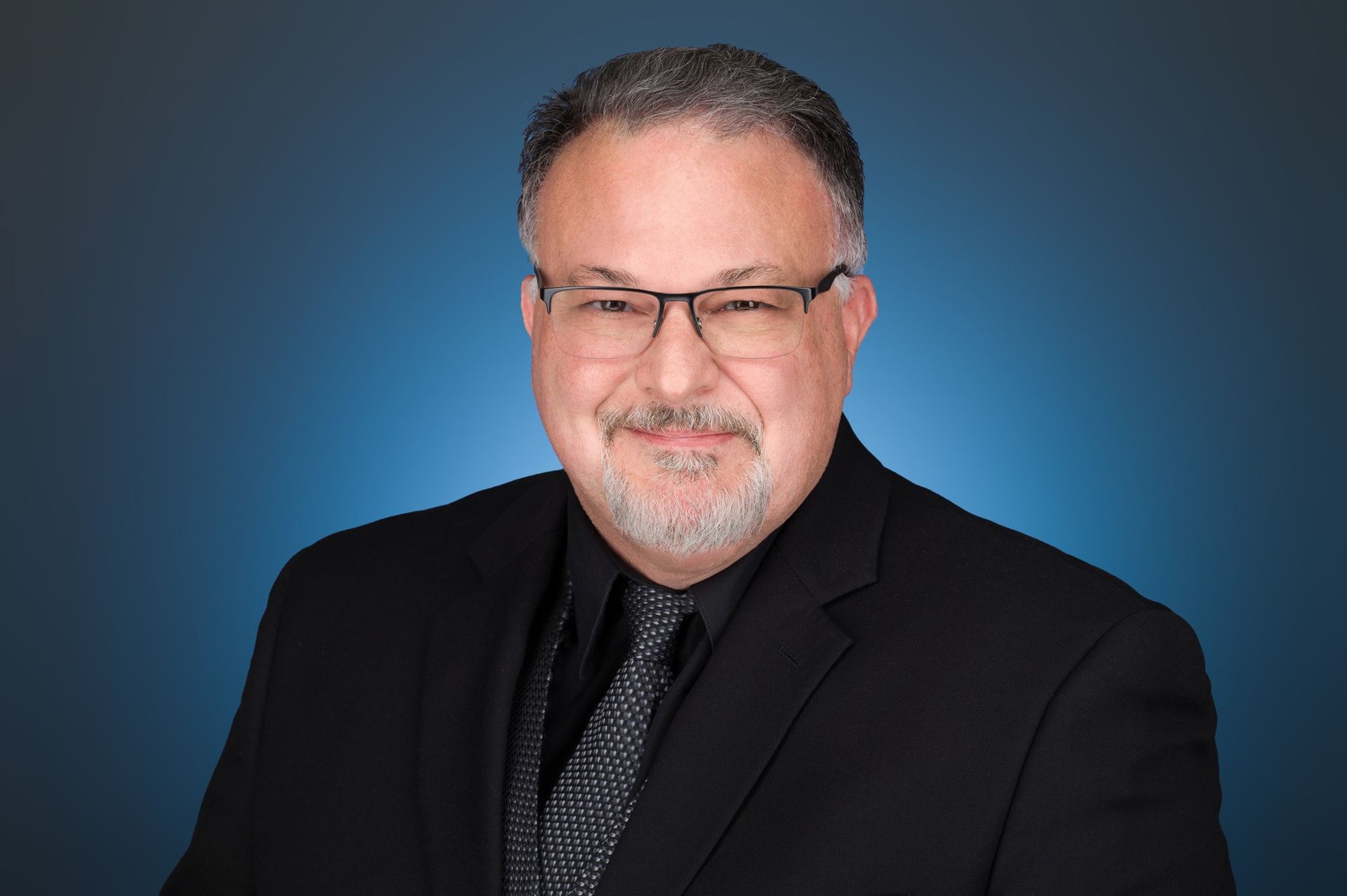 Headshot of a person with glasses, facial hair, and a black suit and tie, against a blue gradient background.