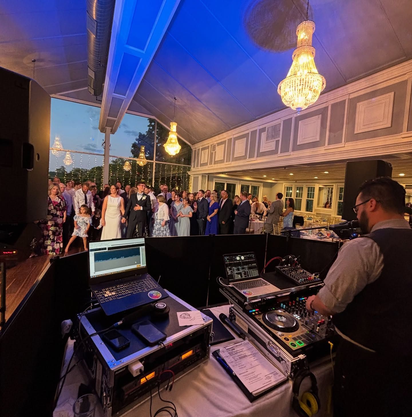 A DJ stands at a console with a laptop and sound equipment, facing a crowd dancing in a lit, elegant event space.