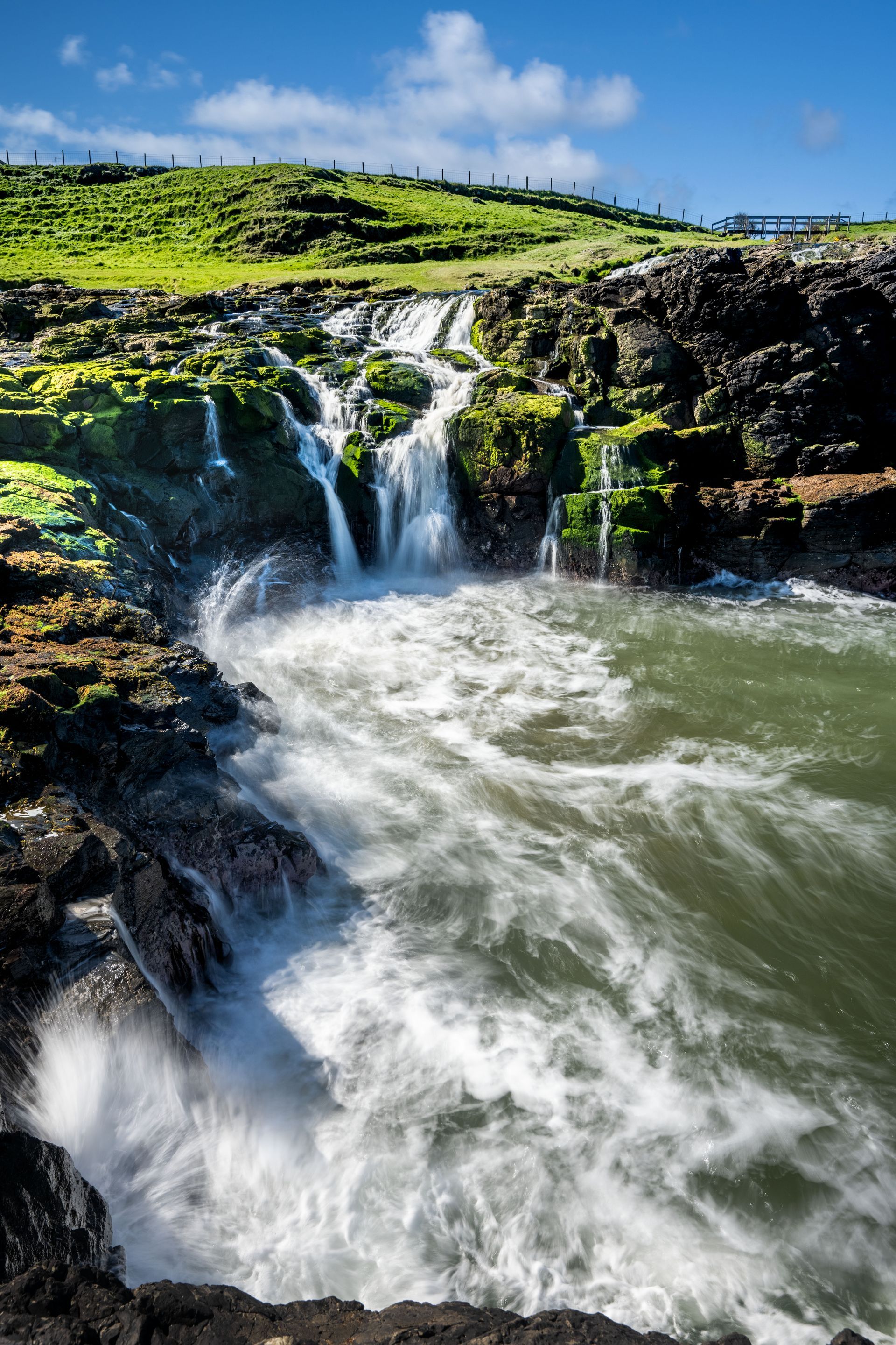 a waterfall is coming down a cliff into the Irish Sea.  Nunseverick Waterfall, Northern Ireland, Talking Excitingly, Brad Burgess Photography
