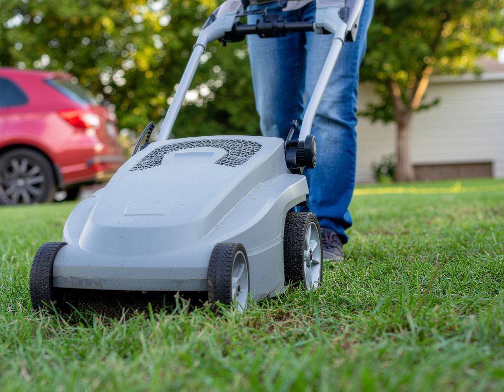 Person mowing a green lawn with a gray push mower; a red car and a house are in the background.