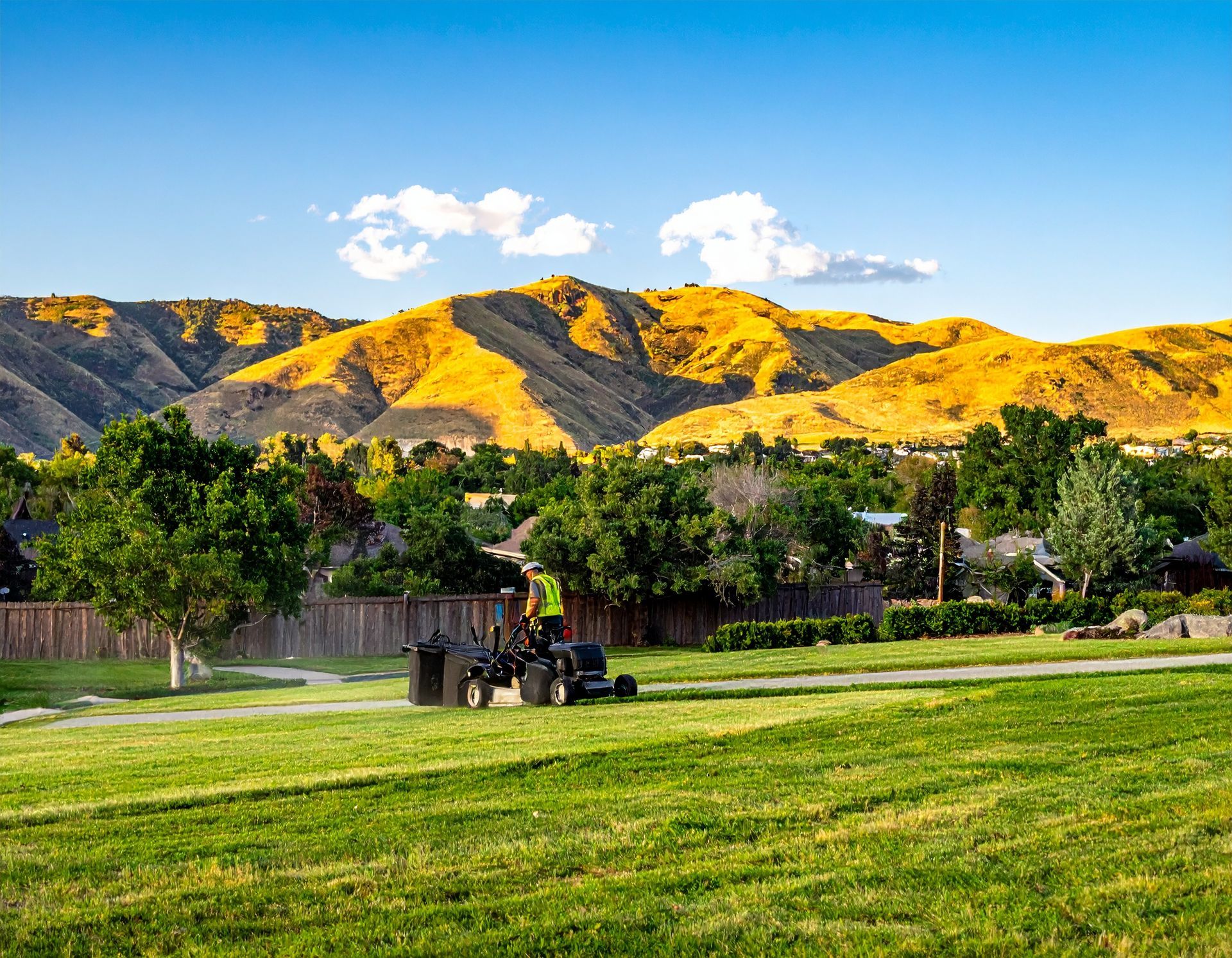 Park lawn with a tractor spraying water, green trees, and sunlit mountains under a blue sky.