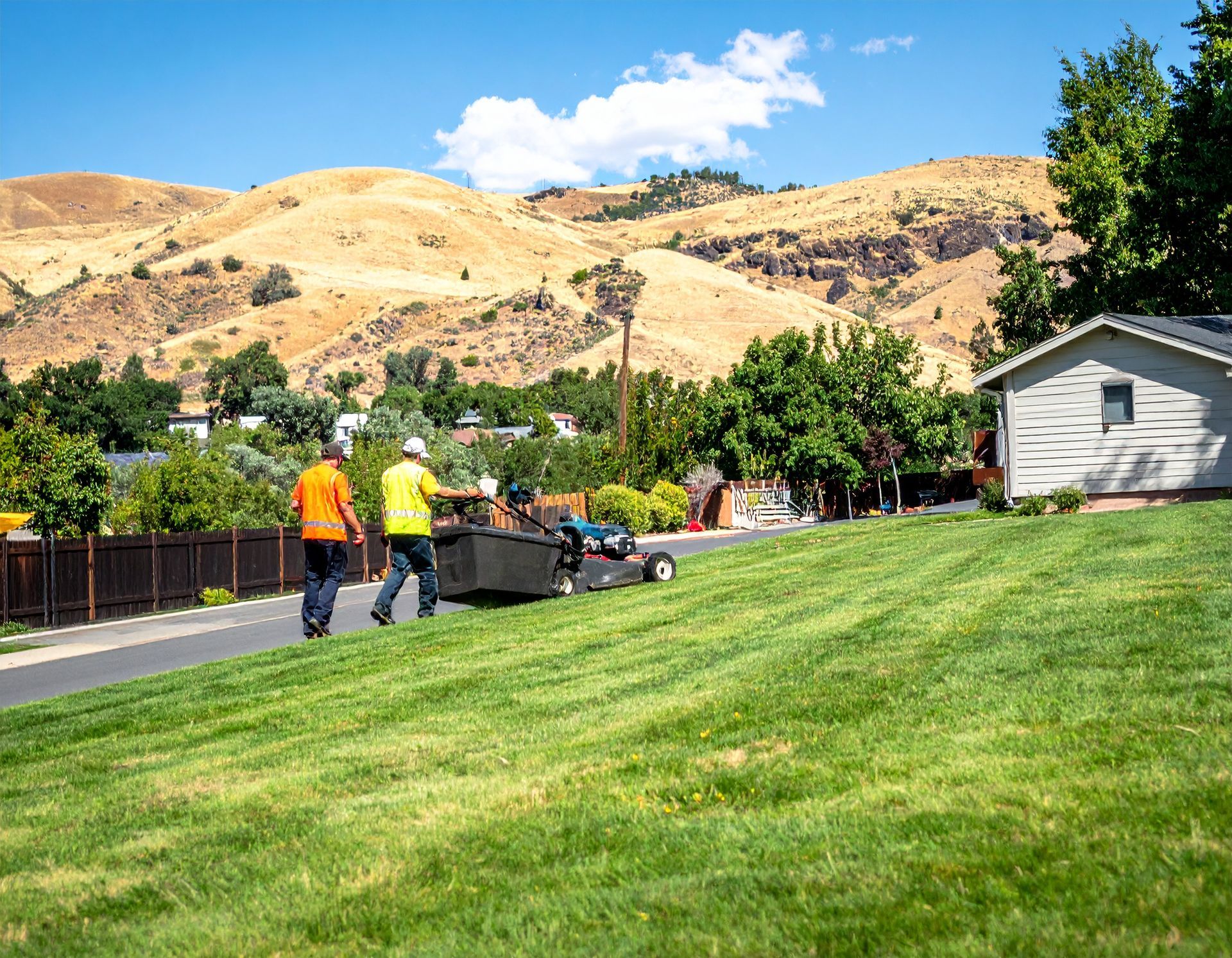 Workers mowing a green lawn near a house, with hills and blue sky in the background
