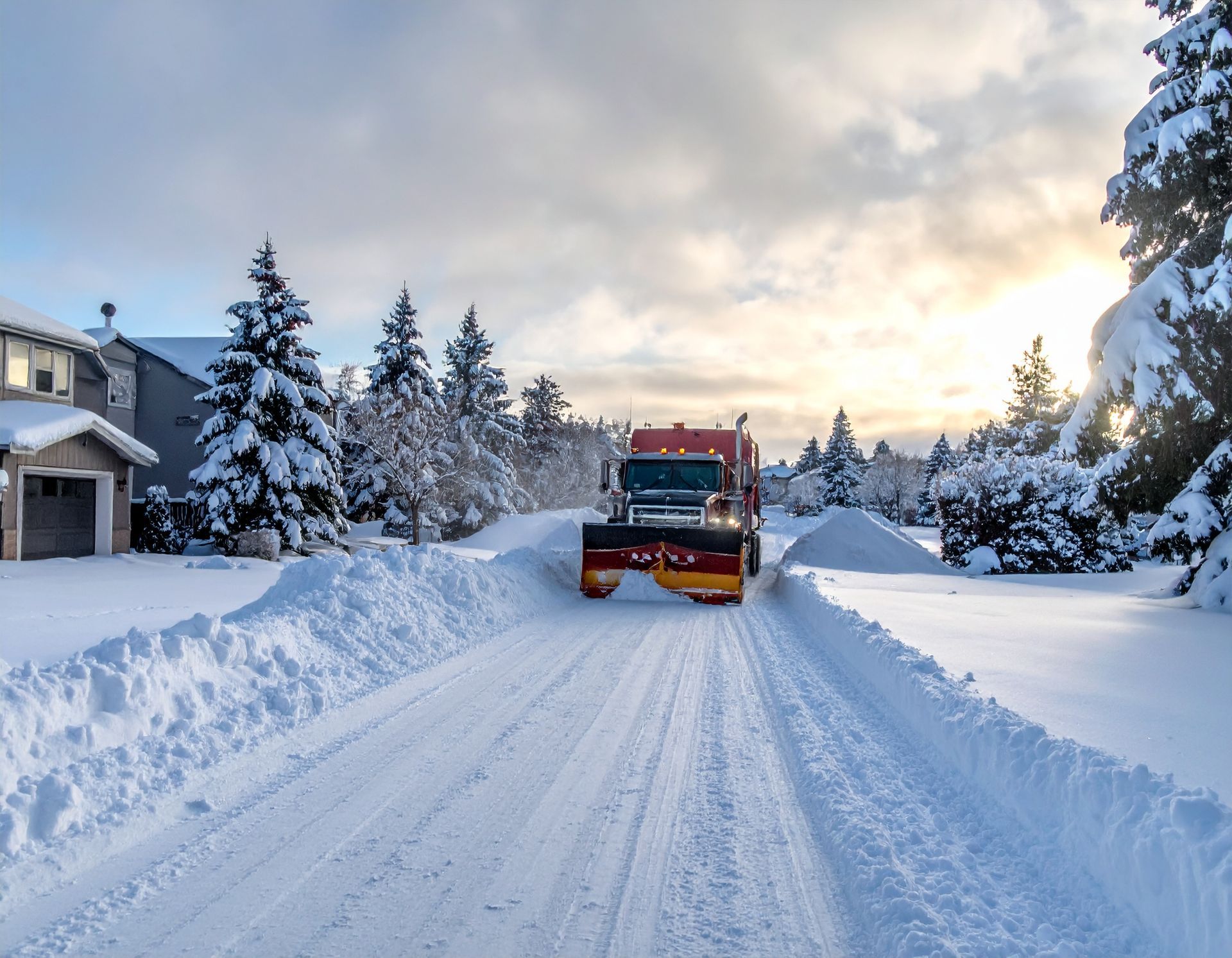 Red snowplow clearing a snow-covered residential street at sunrise, with tall snowbanks on both sides.