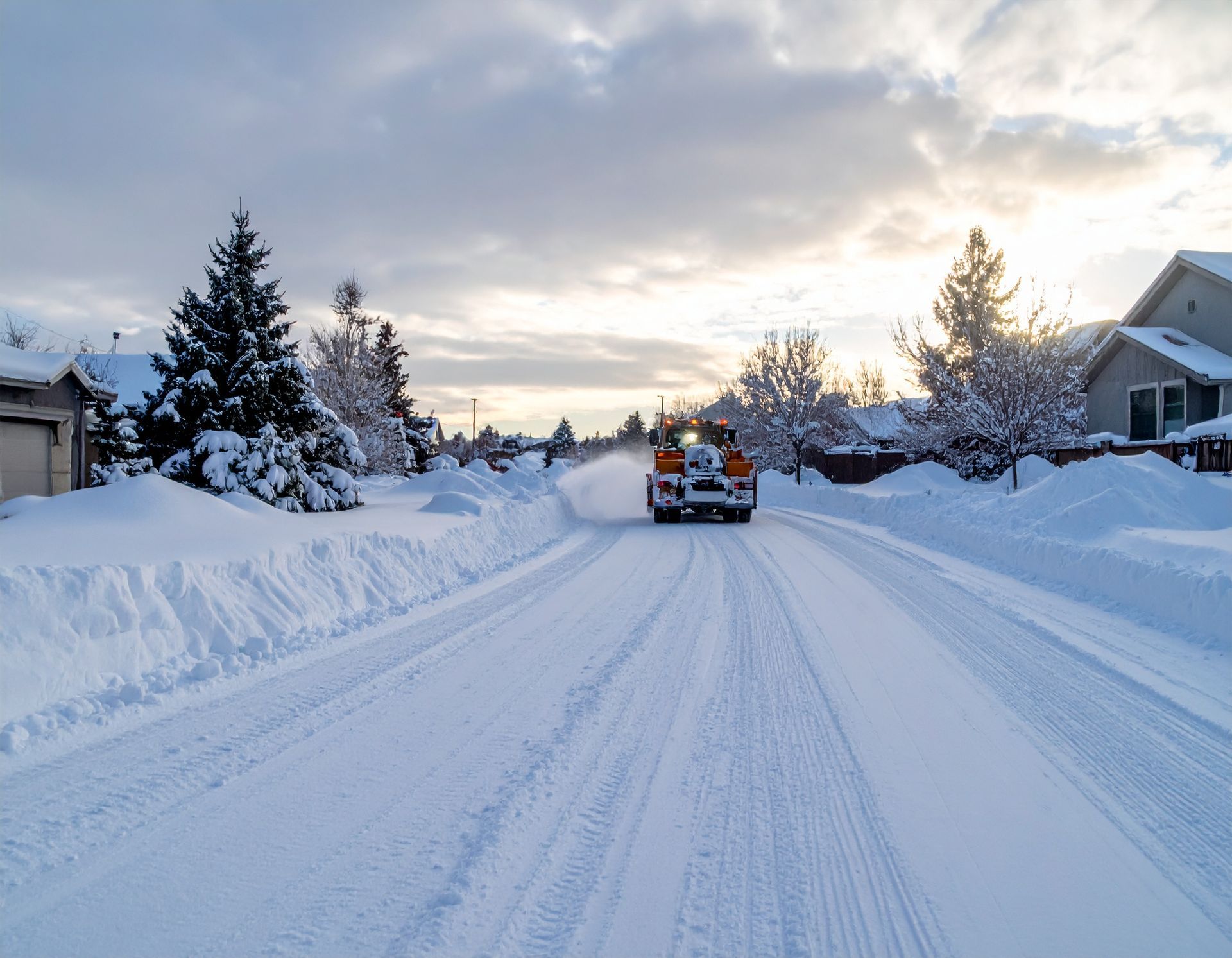 Snow-covered suburban street with a snowplow clearing the road at sunrise.