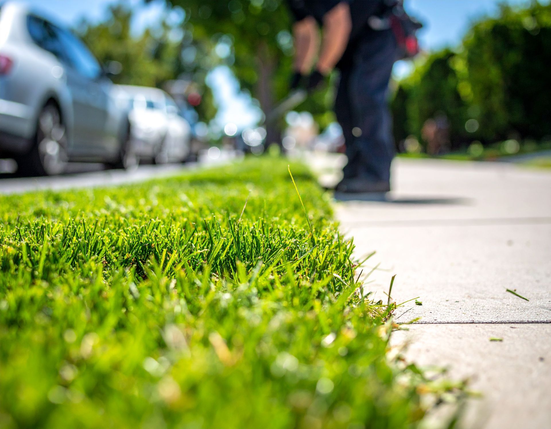 Low-angle view of a sidewalk beside green grass, with a blurred person and parked car in the background