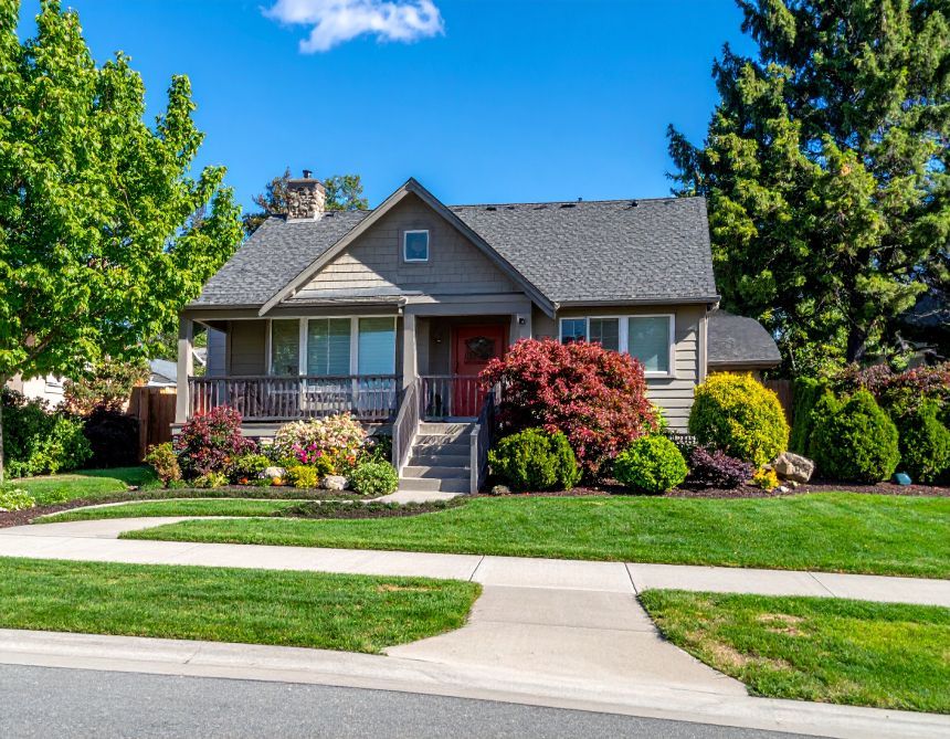 Small, one-story house with gray roof, porch, and colorful landscaping on a sunny day.