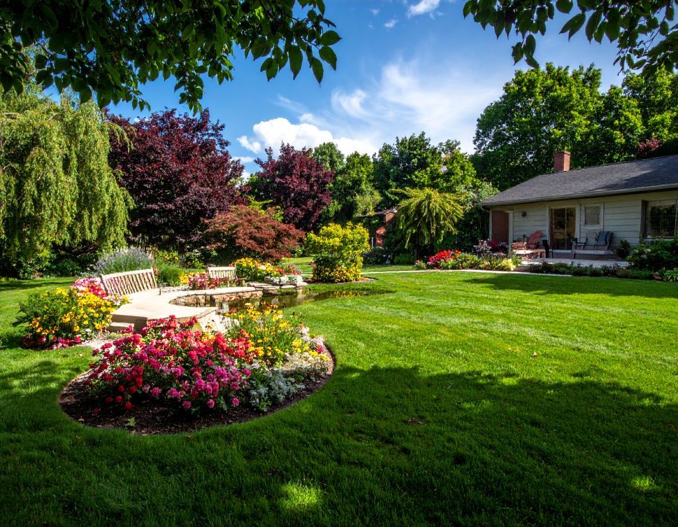 Lush green lawn with colorful flower beds, trees, and a house under a bright blue sky.
