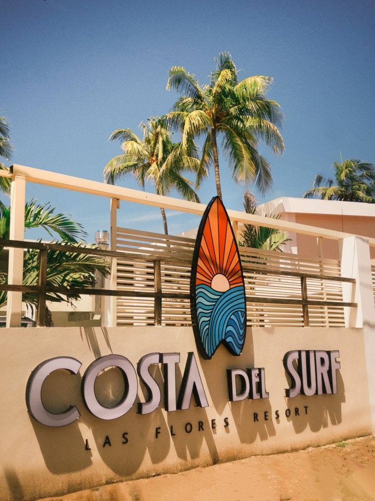 Letrero del resort Costa Del Surf con decoración de tablas de surf, palmeras y cielo azul.