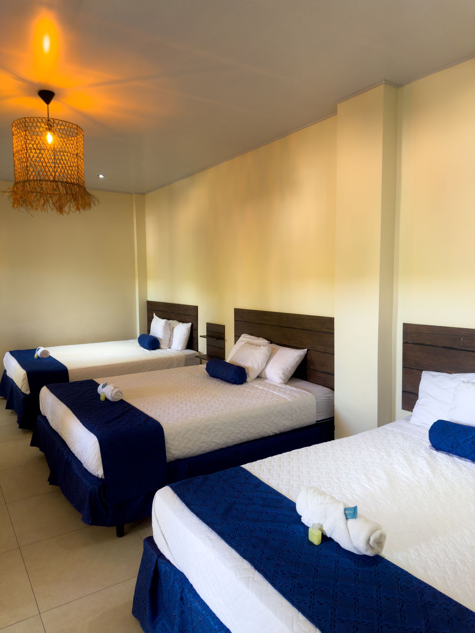 Tres camas en una habitación de hotel con detalles azules, ropa de cama blanca y una lámpara tejida.