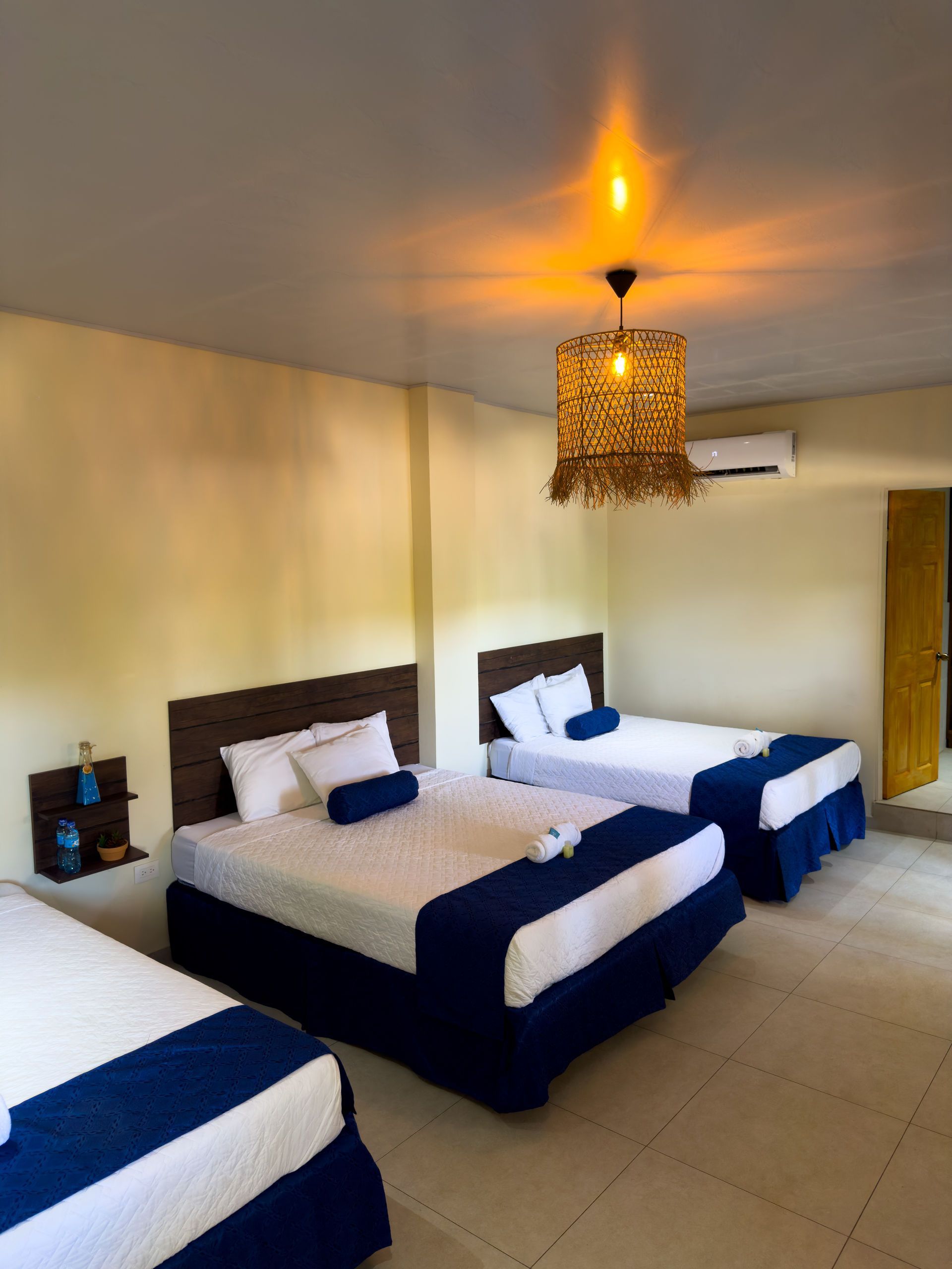 Tres camas en una habitación con sábanas blancas y detalles en azul. Una lámpara de mimbre cuelga del techo.