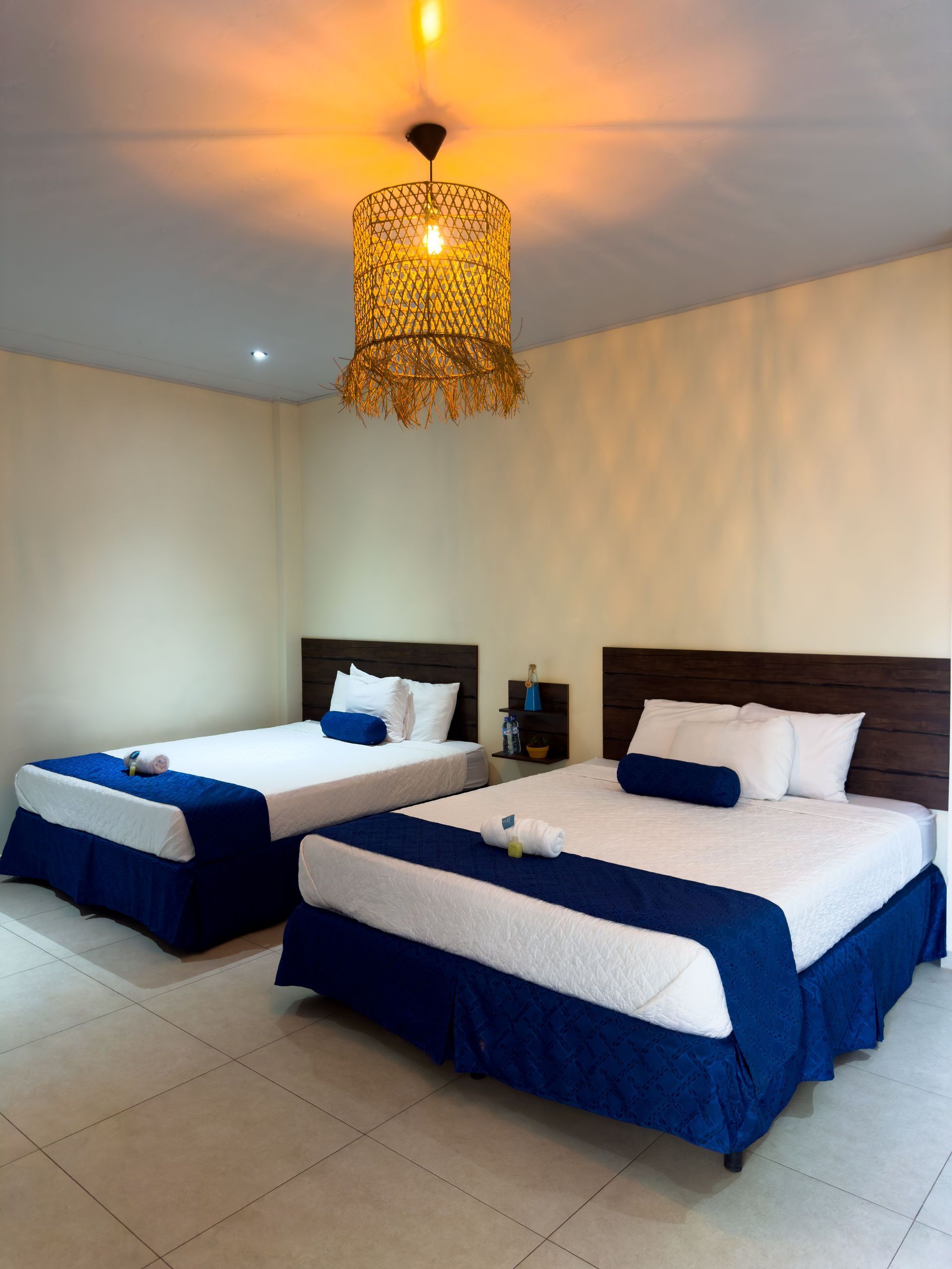 Dos camas en una habitación beige con colchas azules, almohadas blancas y una lámpara decorativa dorada.