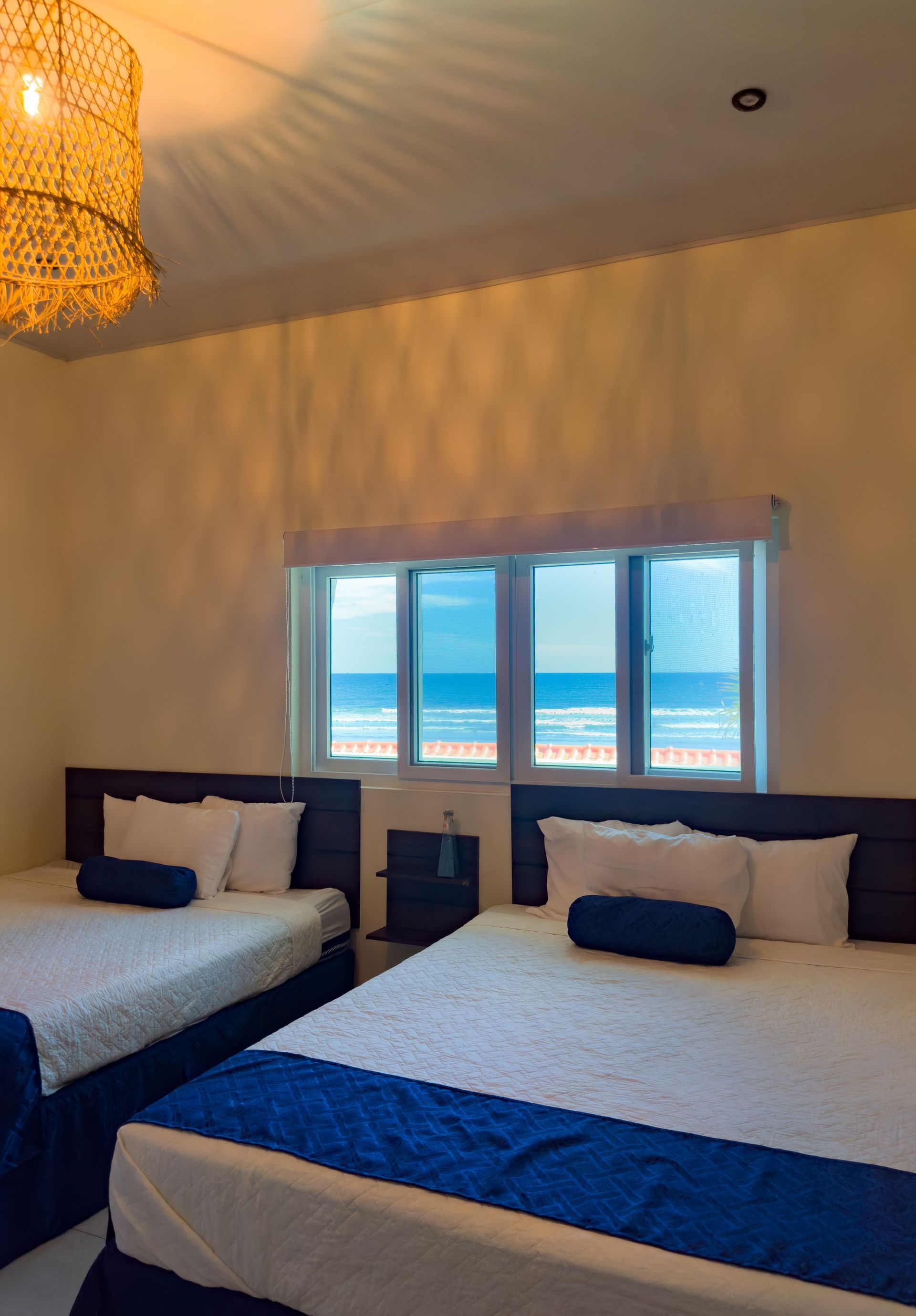 Dos camas queen en una habitación con vista al mar. Cabeceras de madera oscura, ropa de cama blanca y detalles en azul.