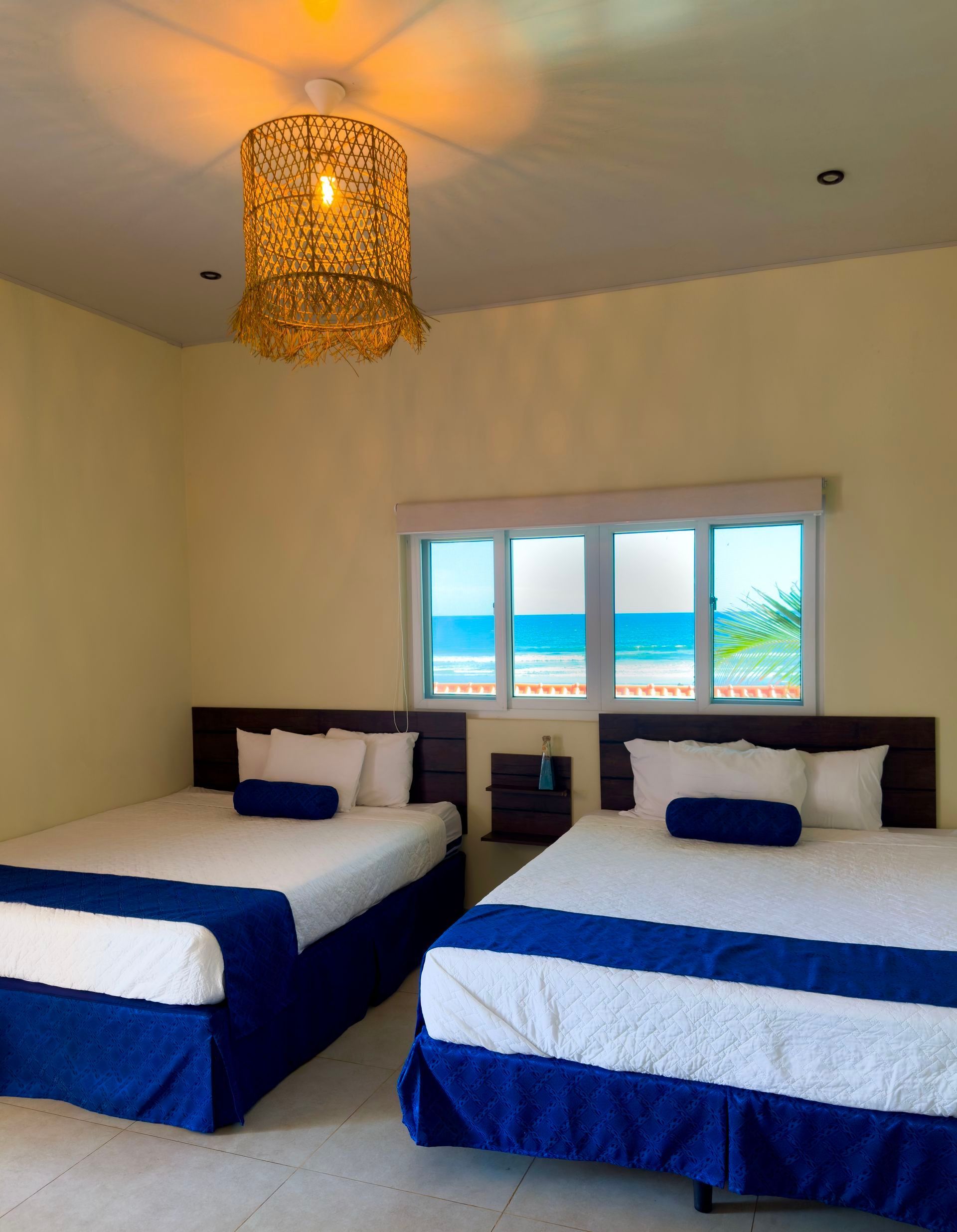 Dos camas en una habitación con vista al mar; ropa de cama azul y blanca, paredes amarillas, lámpara decorativa.