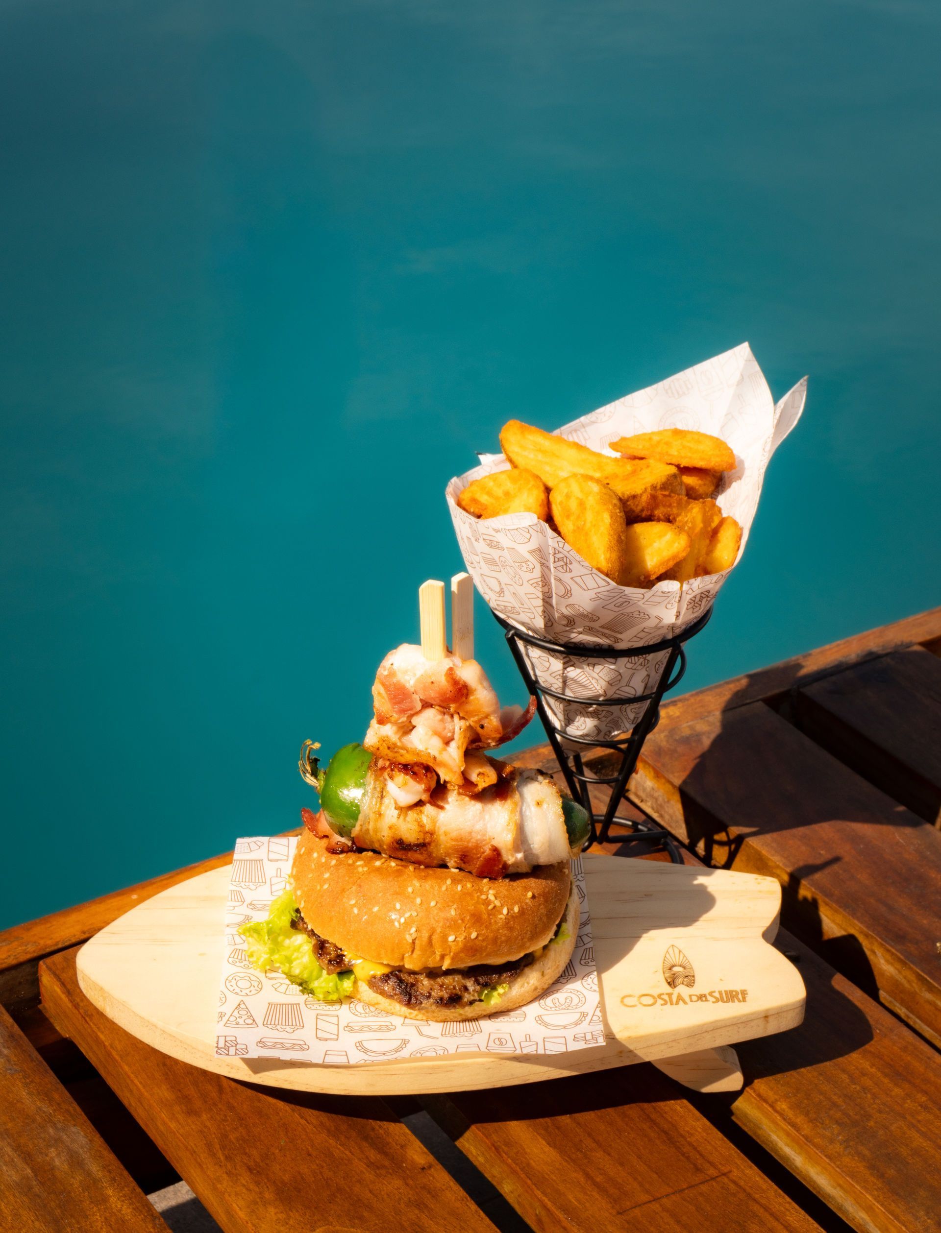 Hamburguesa con patatas fritas en bandeja de madera junto a la piscina.
