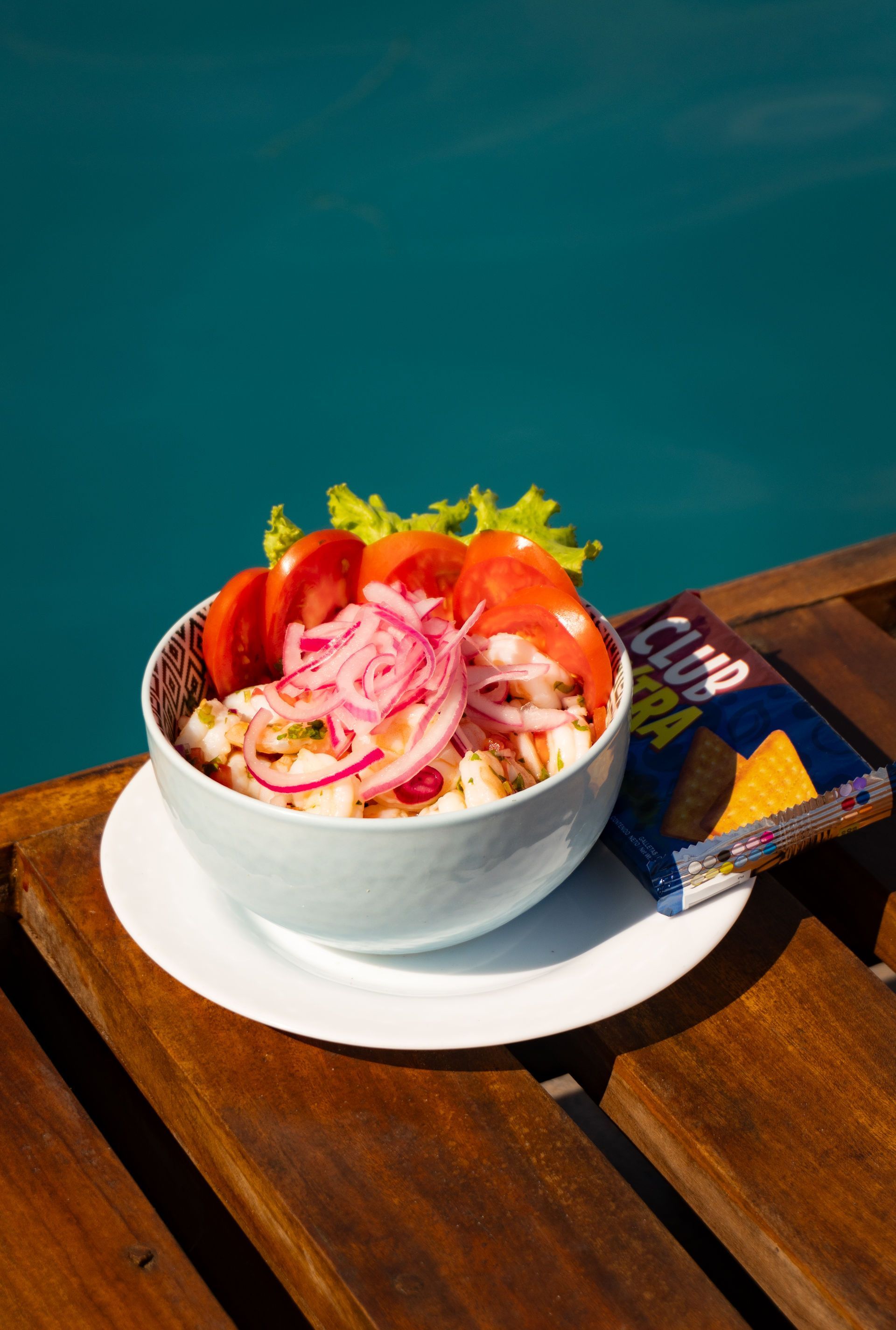 Ceviche en un bowl, cubierto con cebollas rojas y tomates, junto a papas fritas, en un deck de madera con vista a la piscina.