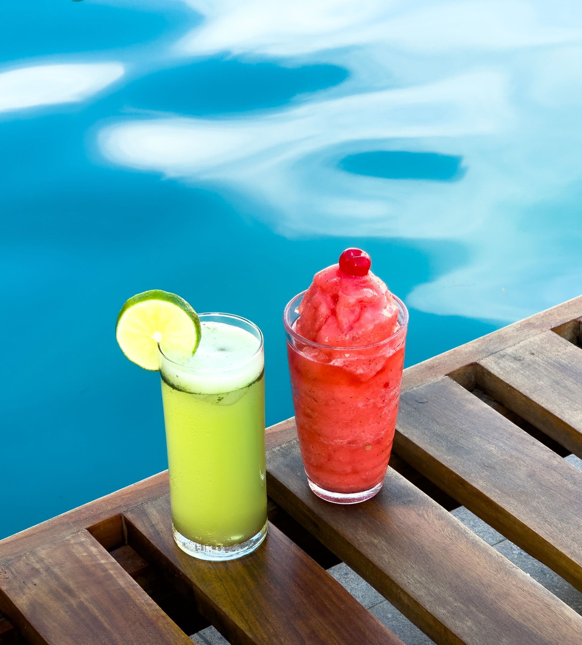 Dos cócteles helados, uno verde con lima y otro rojo con cereza, sobre una superficie de madera cerca de una piscina.