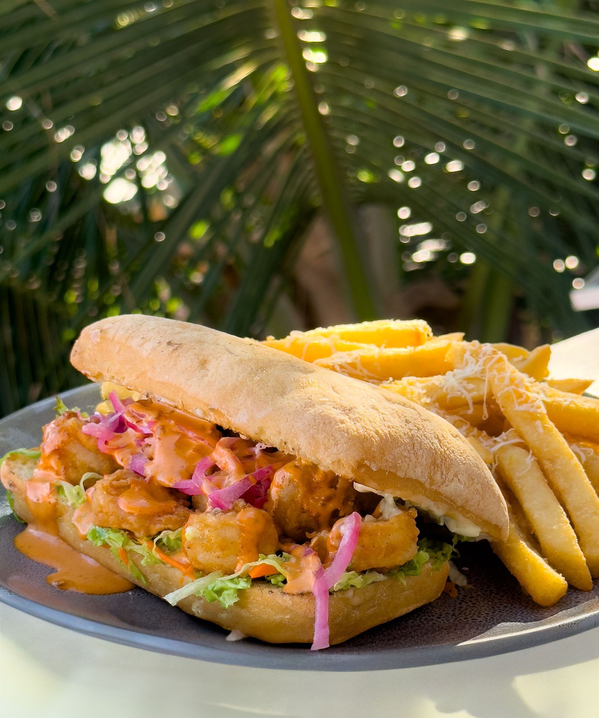 Sándwich de camarones en un panecillo con papas fritas, servido en un plato con hojas de palma de fondo.