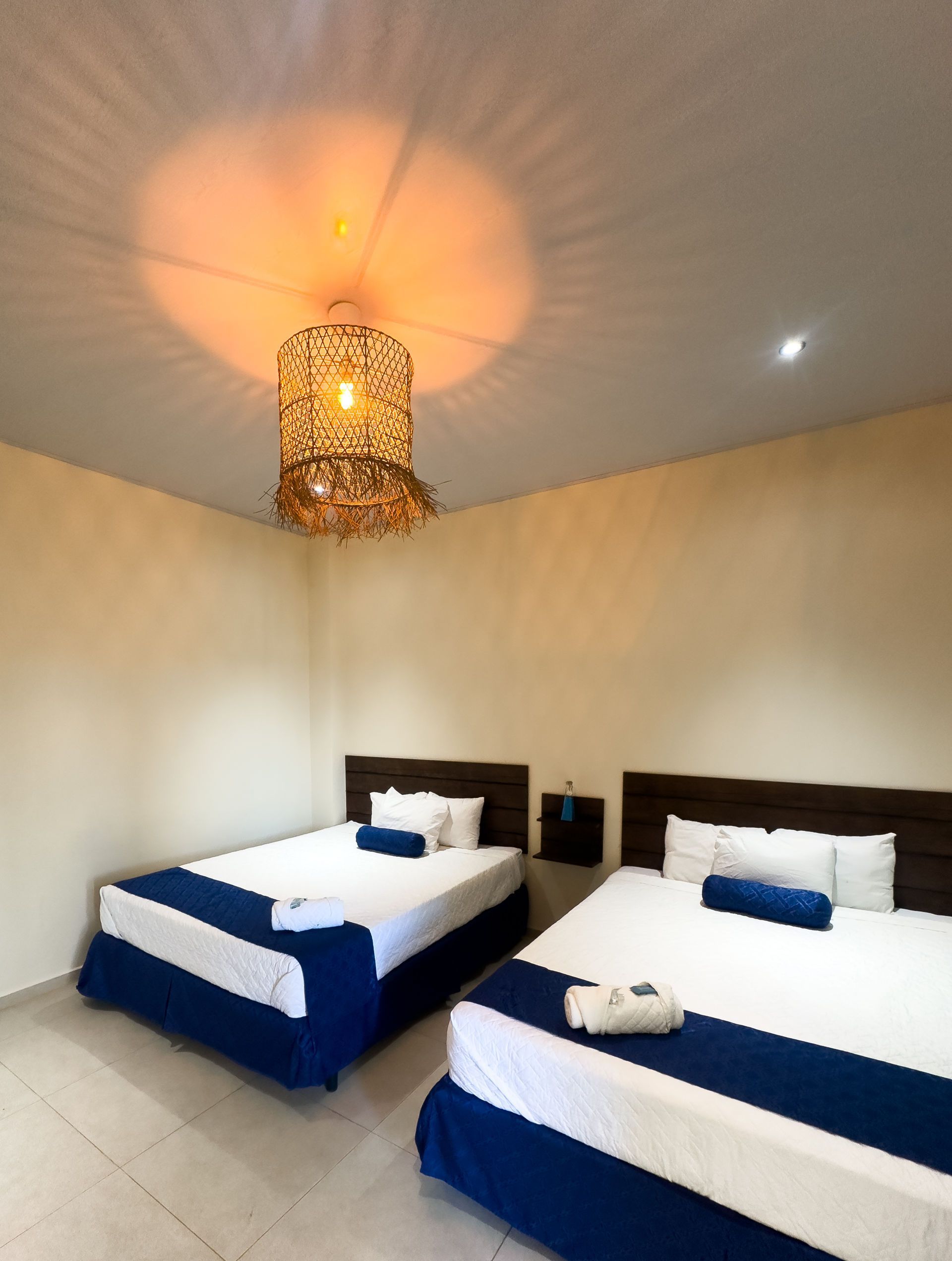 Dos camas con detalles azules en una habitación beige. Una lámpara decorativa cuelga encima.