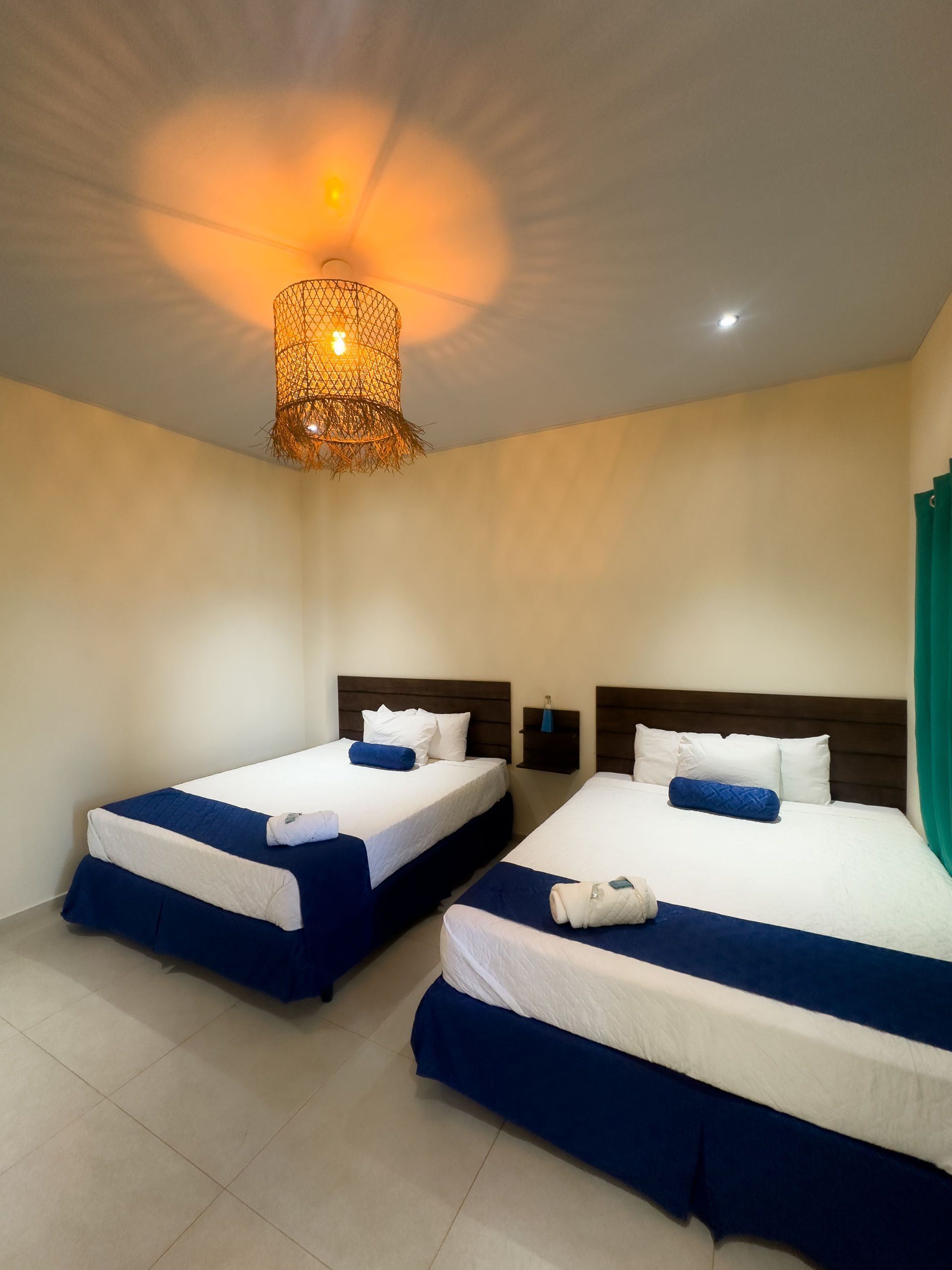 Dos camas con sábanas blancas y detalles en azul en una habitación con una lámpara tejida.