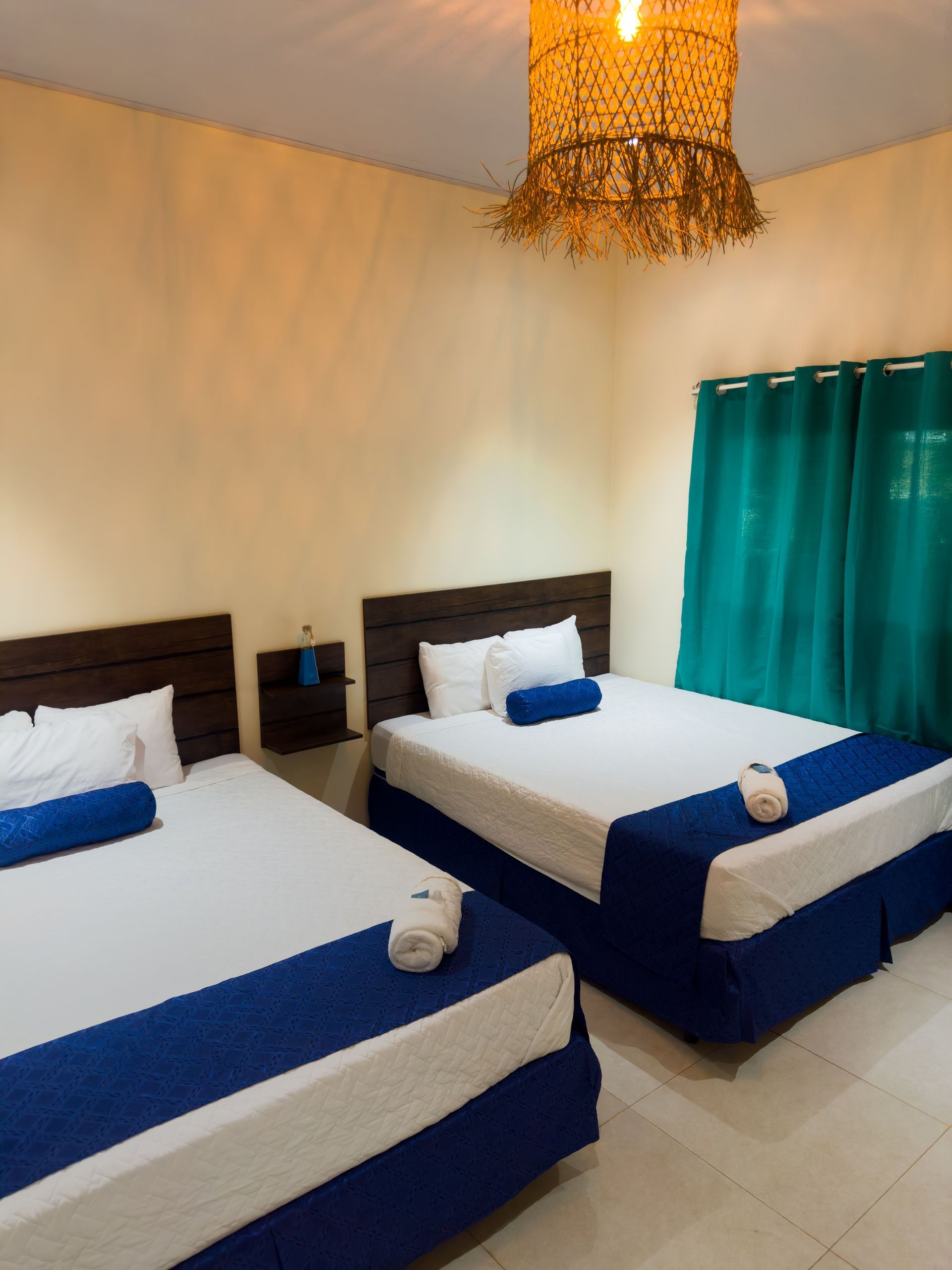 Dos camas con sábanas blancas y detalles azules en una habitación con cortinas verde azulado y una lámpara tejida.