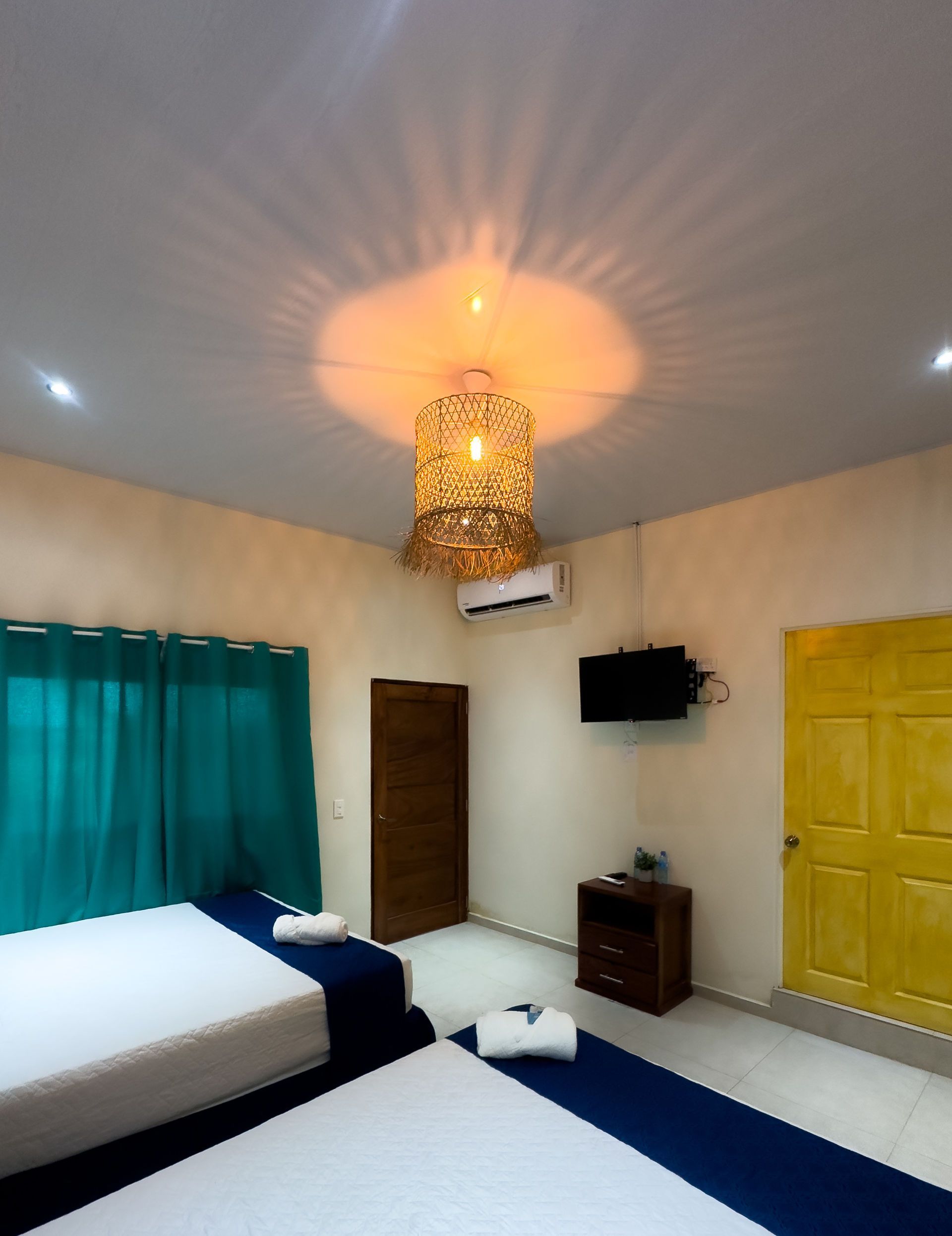 Dormitorio con dos camas, cortinas turquesas, puerta amarilla, lámpara de araña tejida y TV montada en la pared.
