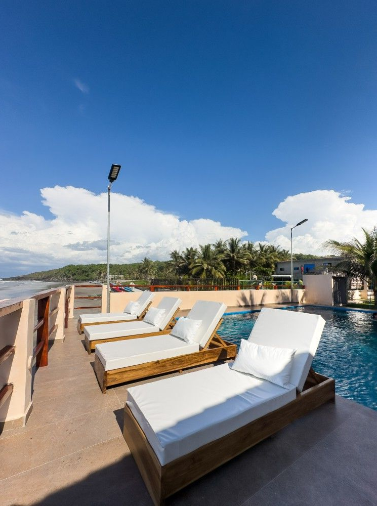 Tumbonas junto a la piscina con cojines blancos bajo un cielo azul brillante con esponjosas nubes blancas.