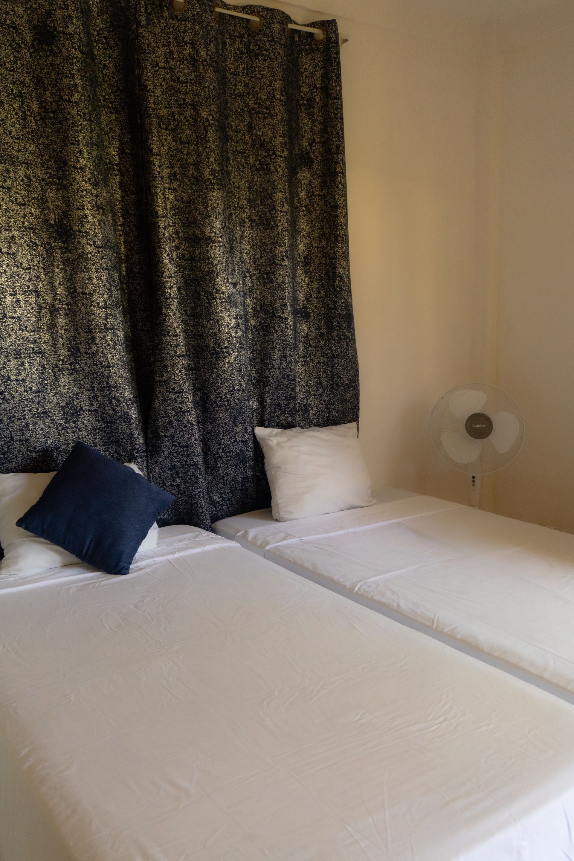 Dos camas individuales con sábanas blancas y almohadas azul marino y blancas; cortinas estampadas negras, un ventilador.