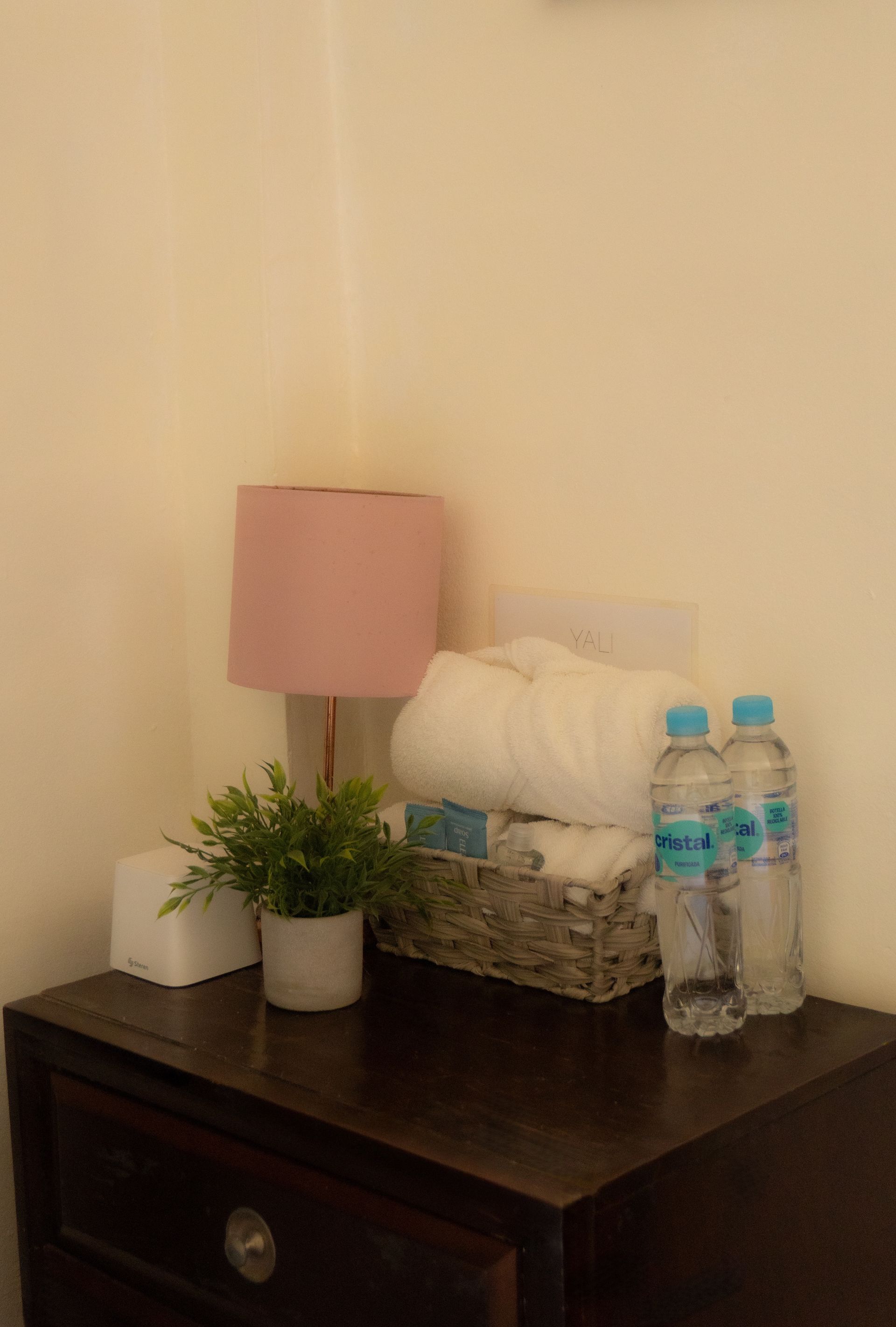 Una mesita de noche de madera oscura con una lámpara rosa, una planta, toallas y agua embotellada en un rincón.