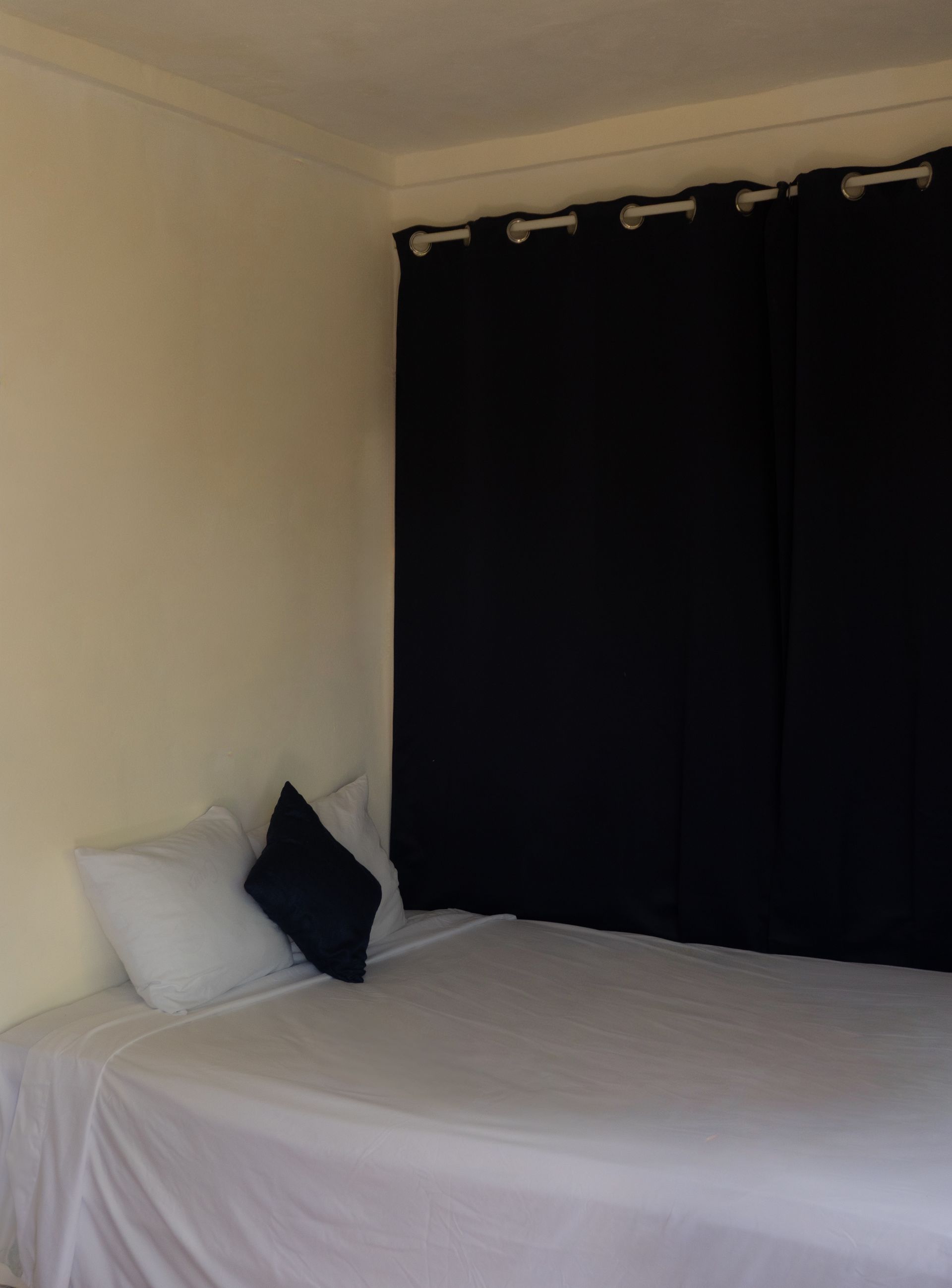 Cama con sábanas y almohadas blancas, almohada decorativa azul marino y cortinas negras. Paredes beige.