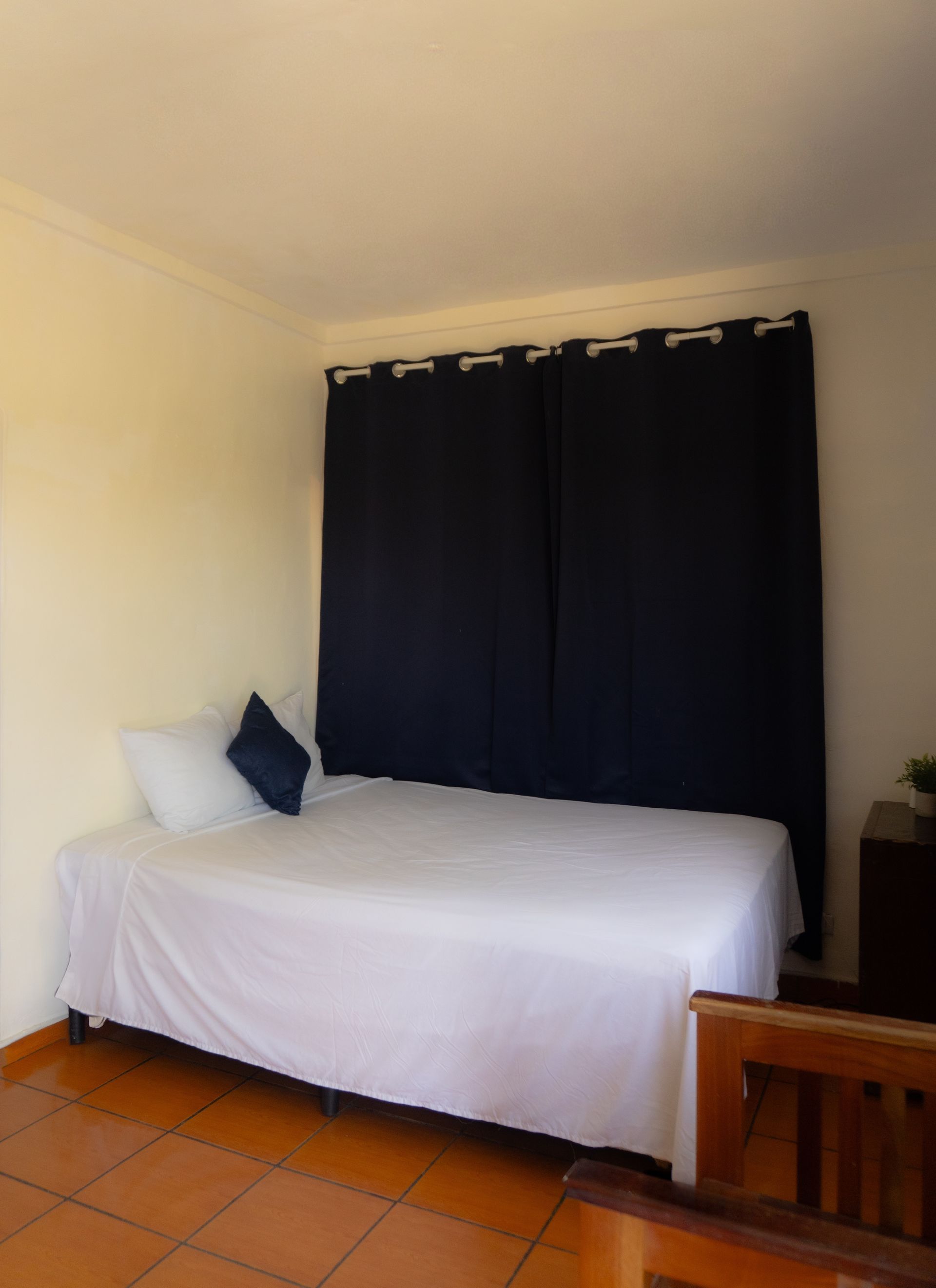 Sofá cama con sábanas blancas, almohadas azules y cortinas azul marino en una habitación con suelo de baldosas.