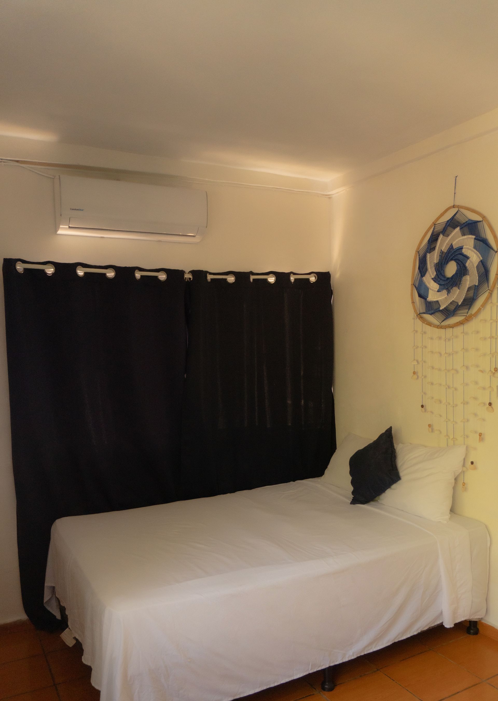 Dormitorio con cama blanca, cortinas negras, atrapasueños y aire acondicionado.