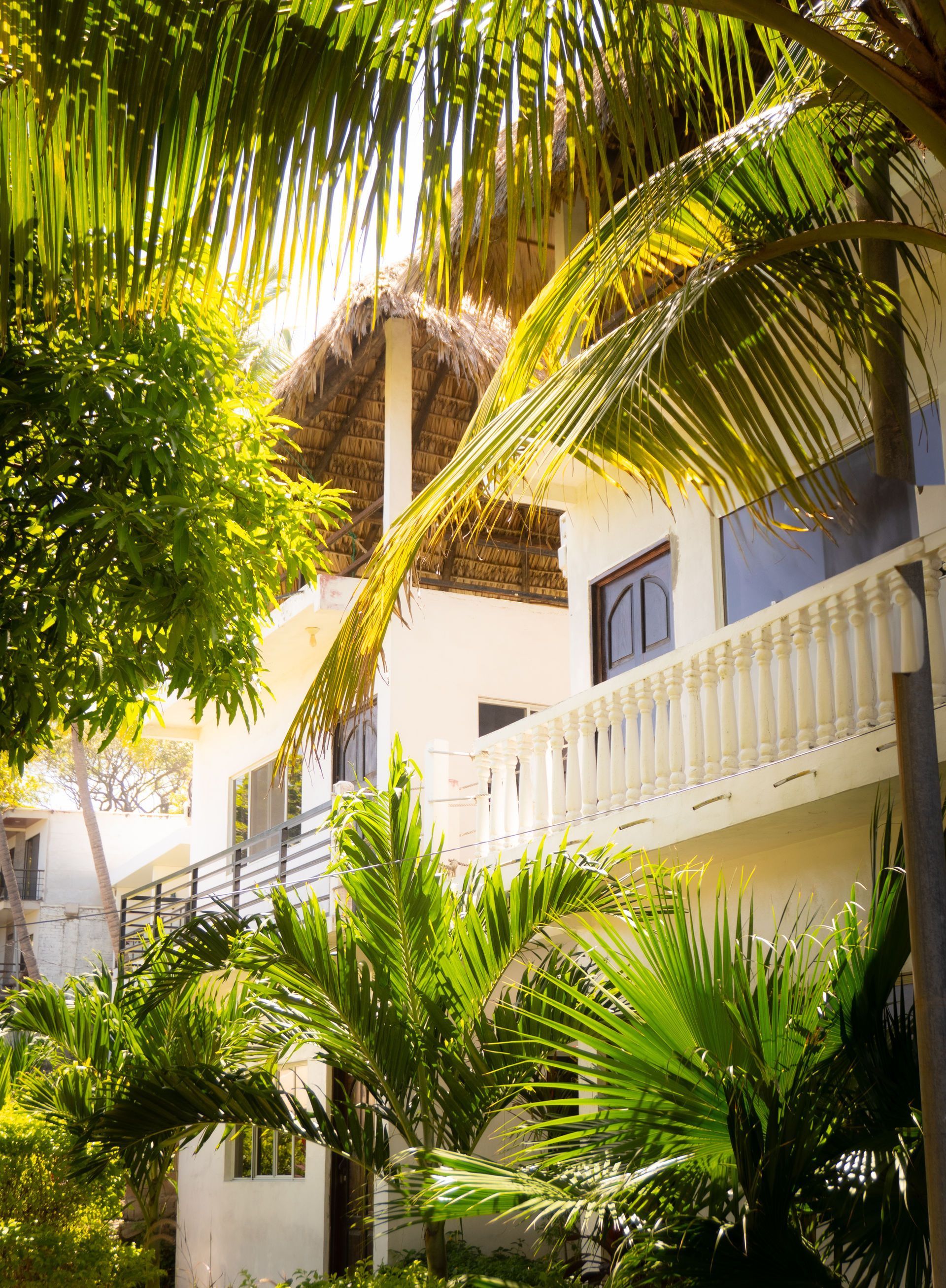 Edificio blanco con balcón, techo de paja, palmeras, luz solar brillante.