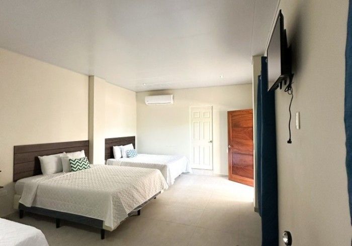 Amplia habitación de hotel con tres camas, televisor y puerta abierta. Paredes beige y ropa de cama blanca.